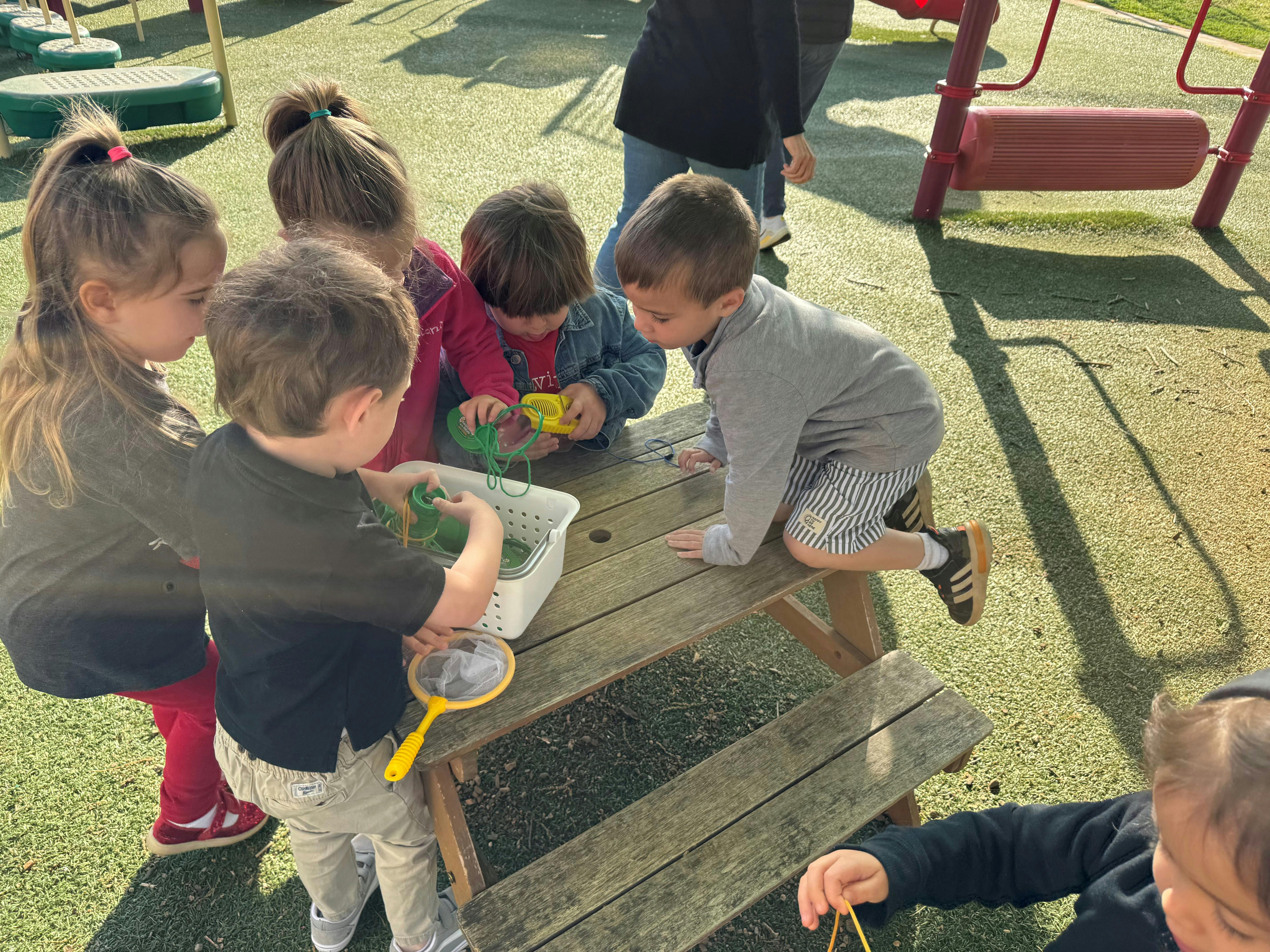 Group of children playing with toy bugs and nets at a small table outdoors