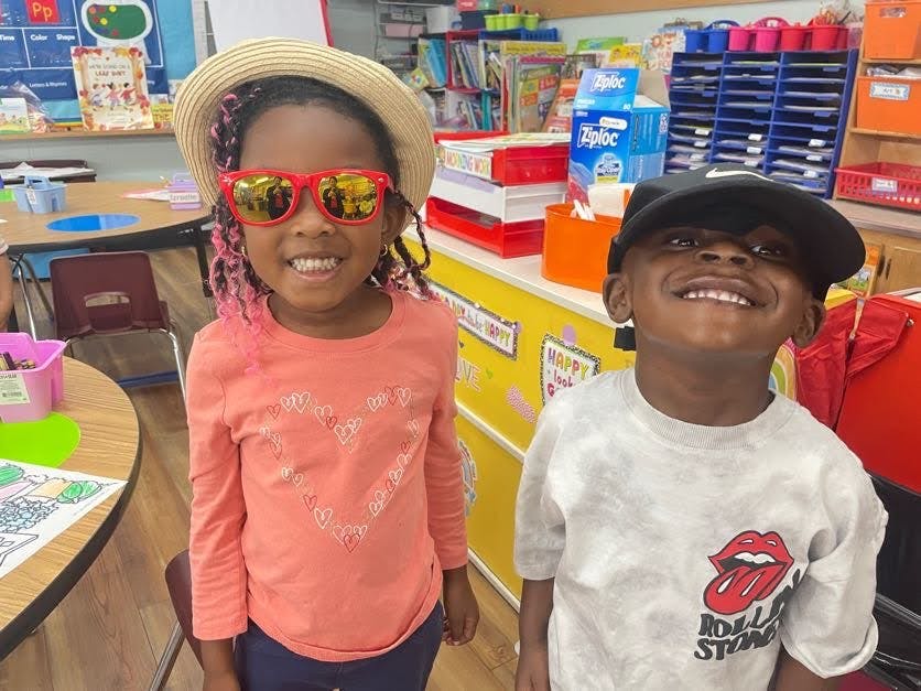 Two children smiling, one wearing a hat and sunglasses, in a classroom