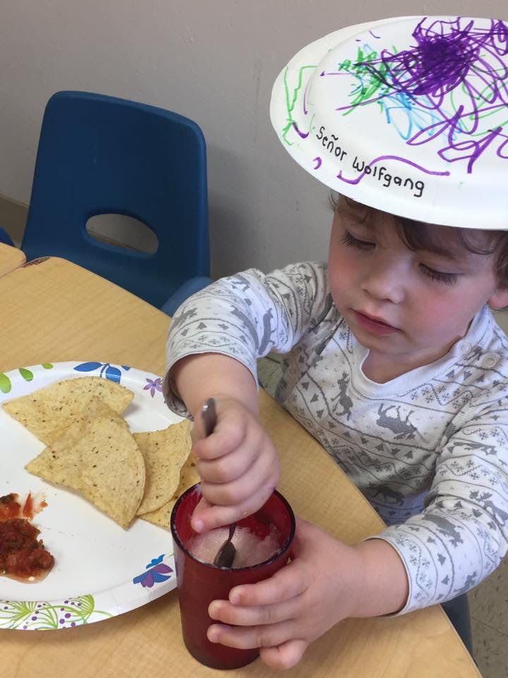 Child wearing a paper plate hat, stirring a cup with a spoon