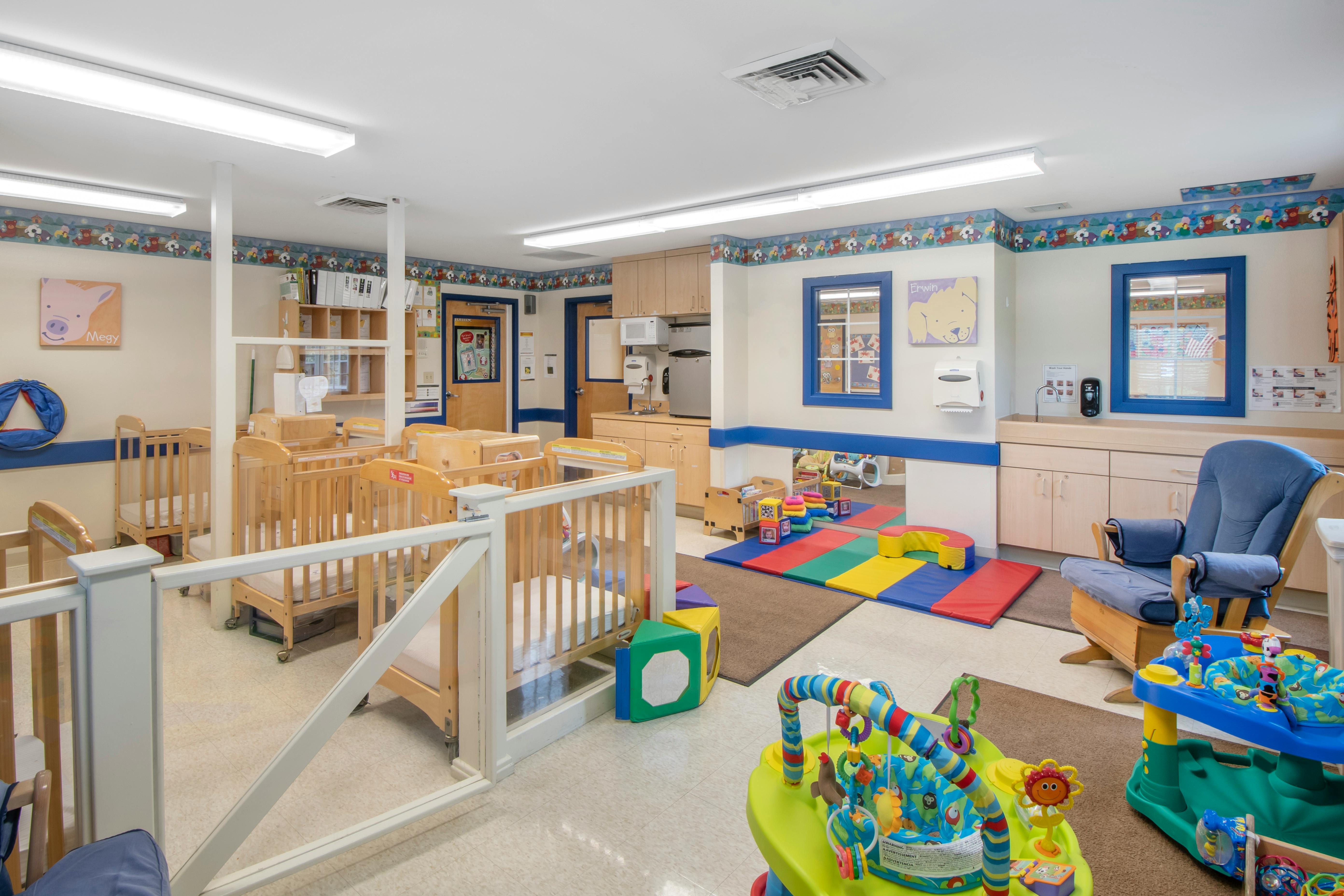A brightly lit room with several cribs, playmats, a rocking chair, and a changing station