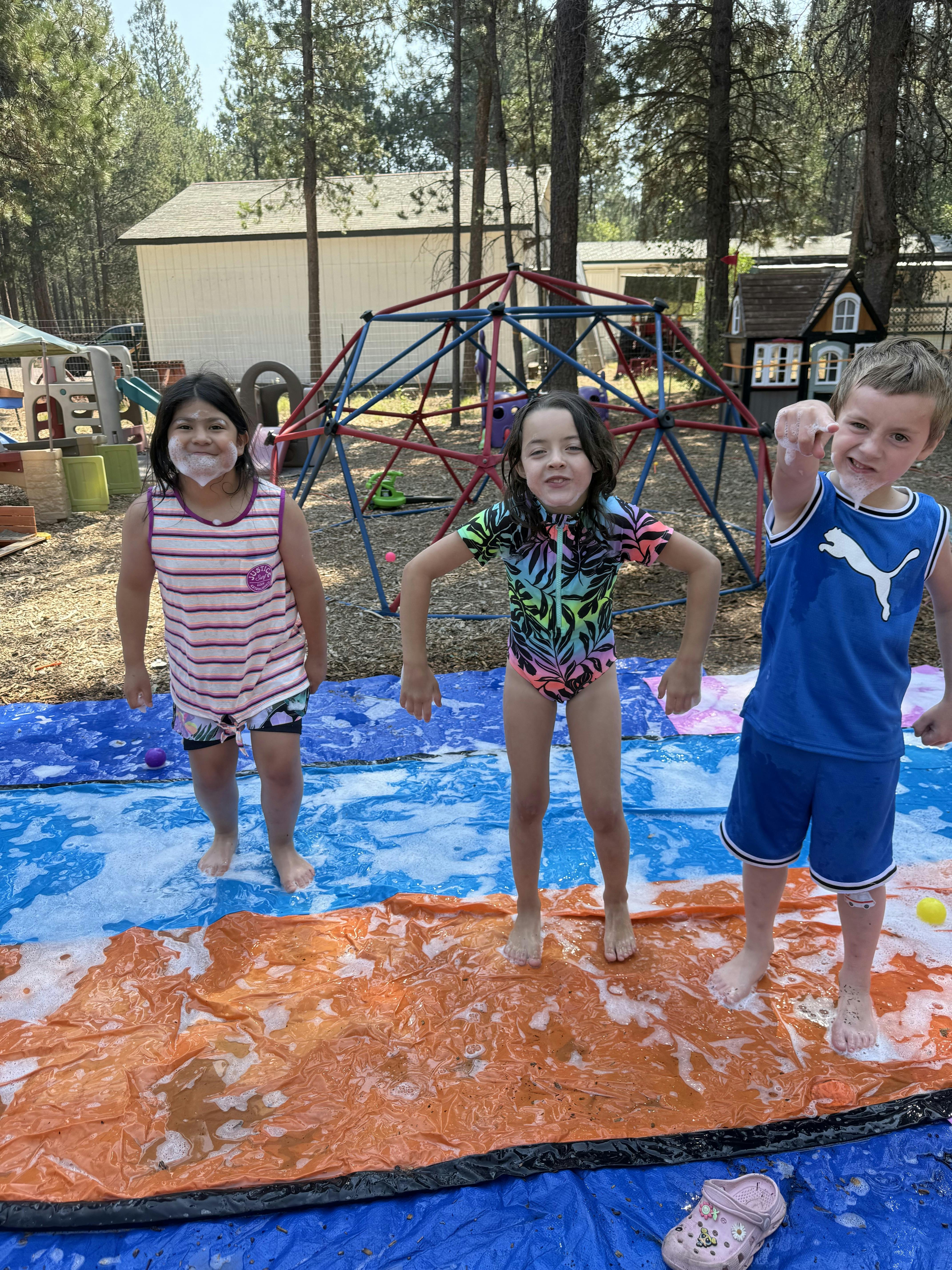 Three children standing on a wet blue and orange tarp outdoors