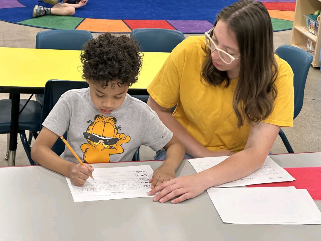 Person helping a child write on paper at a table