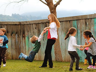 Adult holding a child by the hands, swinging them in a yard with other children nearby