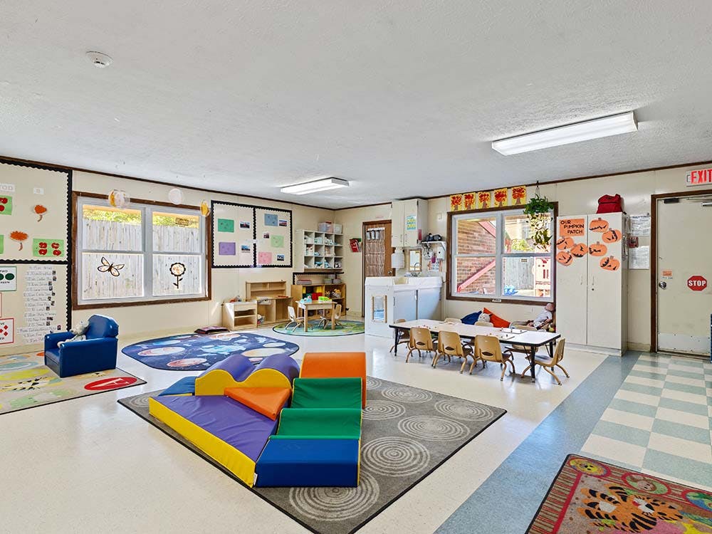 An empty childcare classroom with play mats, small tables, chairs, and windows