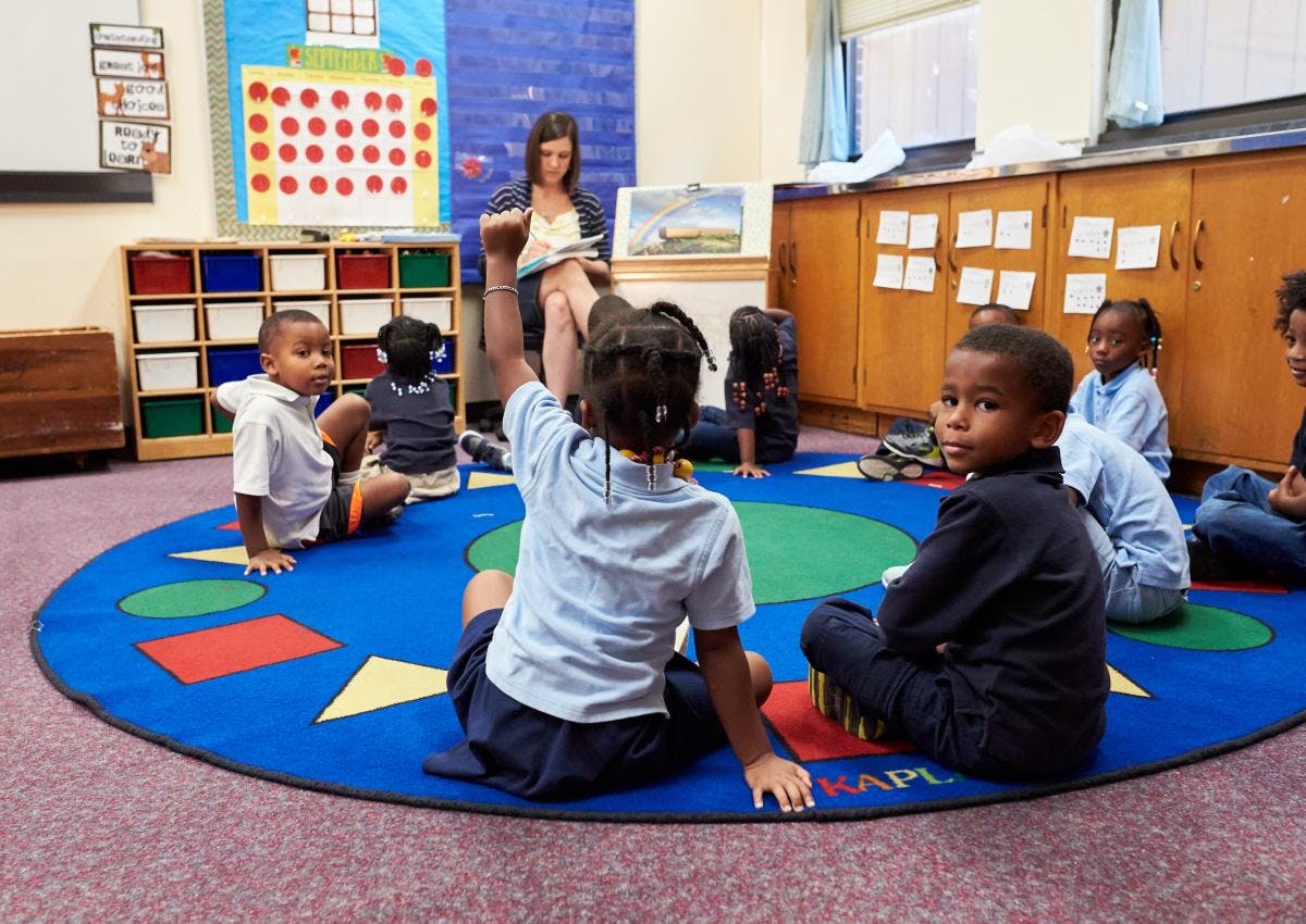 Children sitting on a rug in a classroom with one child raising a hand