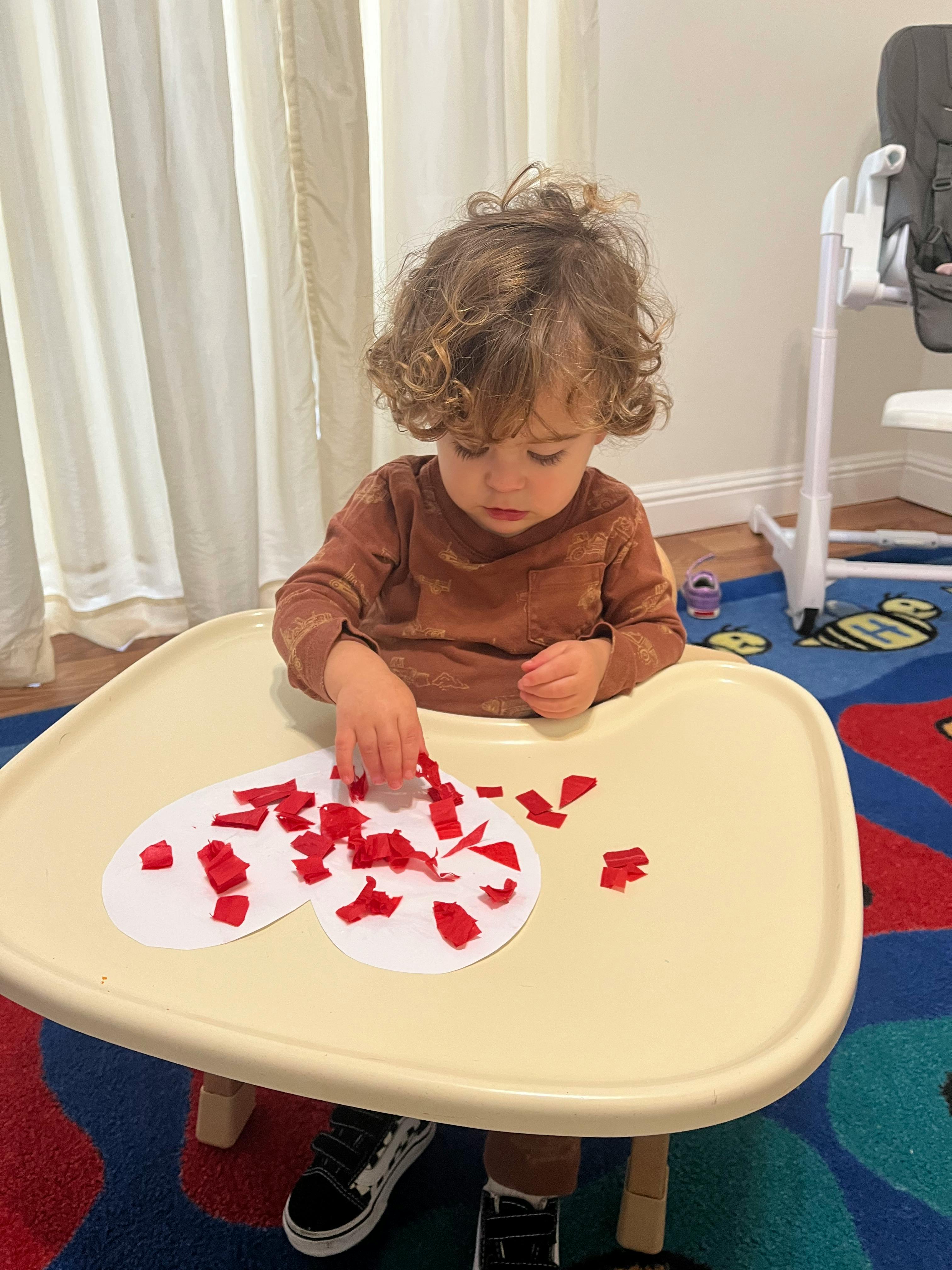 Child sitting in a high chair, placing red paper pieces on a heart shape