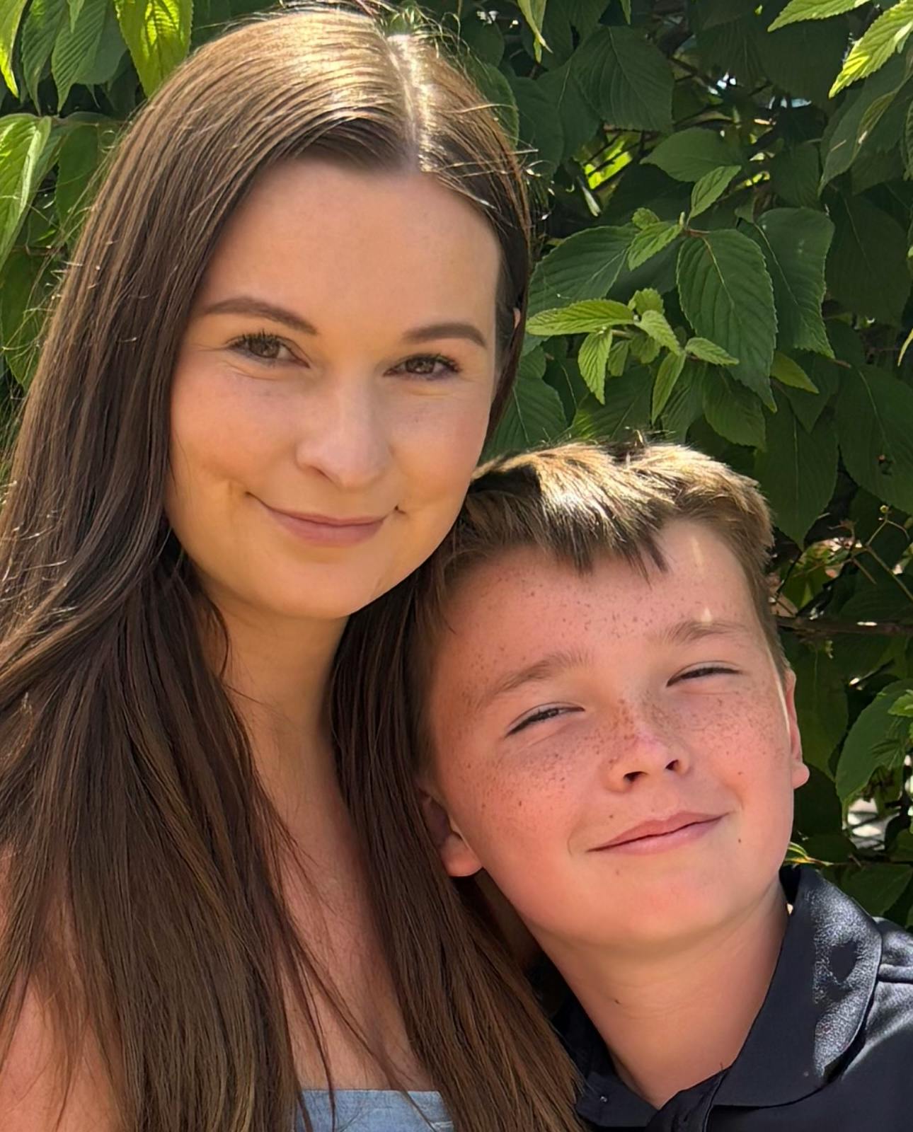 A person and a child posed together outdoors in front of foliage