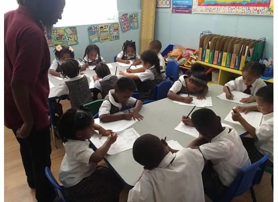 Person standing over children writing at tables in a classroom