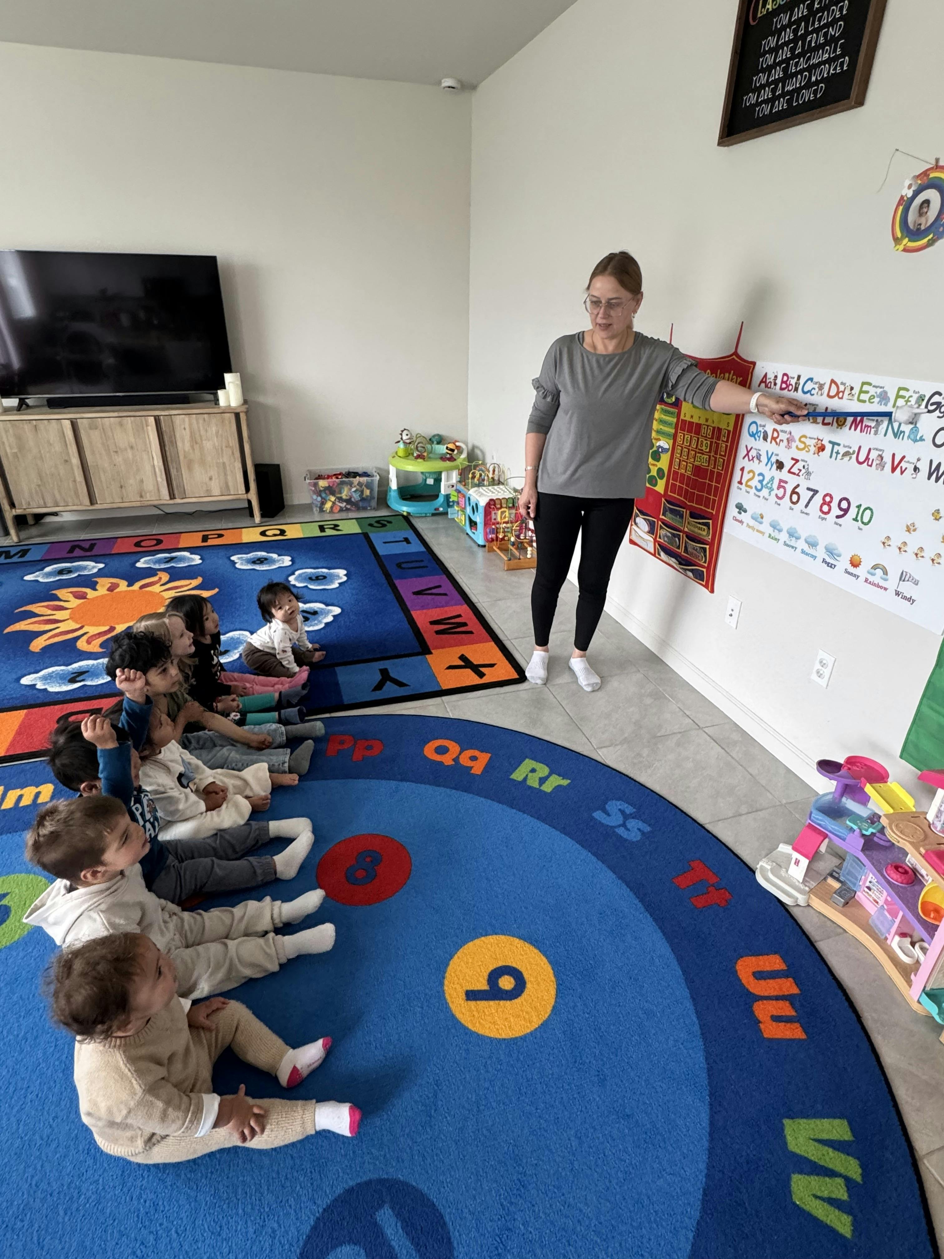 Adult pointing to an alphabet chart for a group of children sitting on a rug