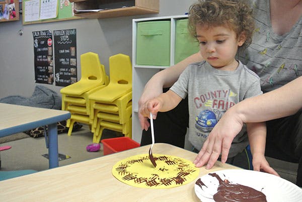 Child decorating a yellow paper plate with chocolate using a spoon and brown substance