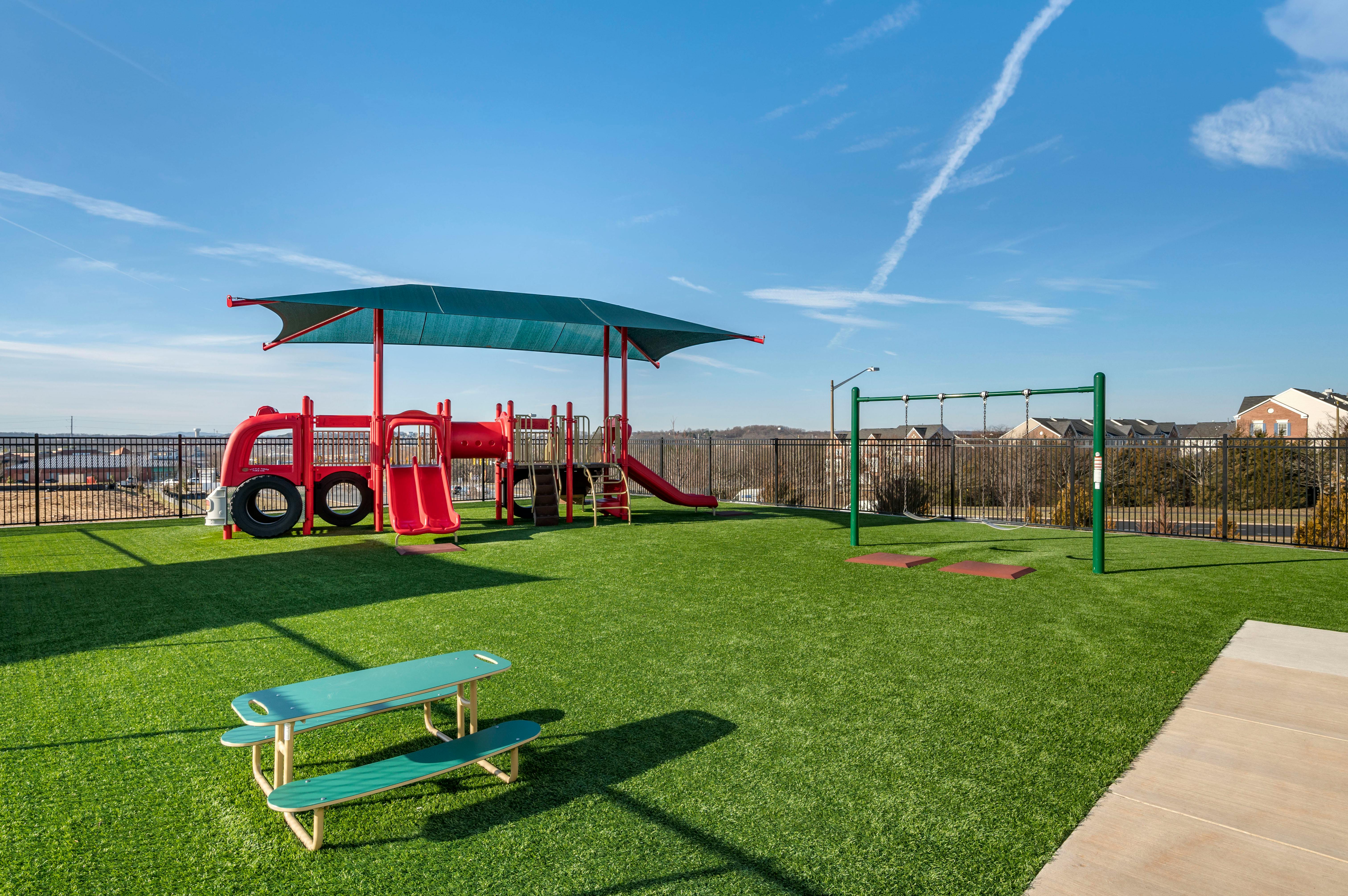 A fenced playground with green artificial turf, a red bus-shaped play structure, and swings