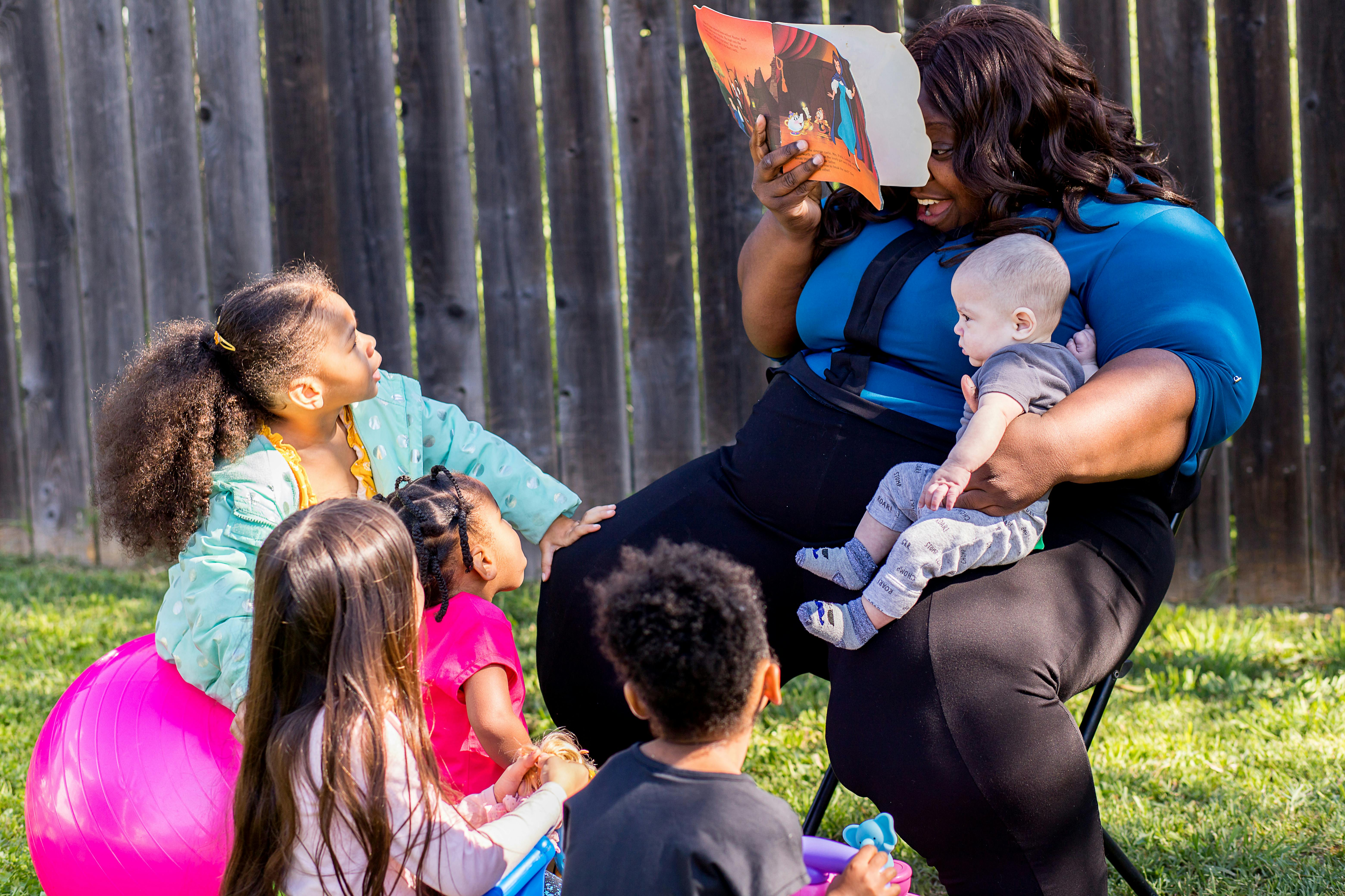 Person reading a book to a baby and several children outdoors