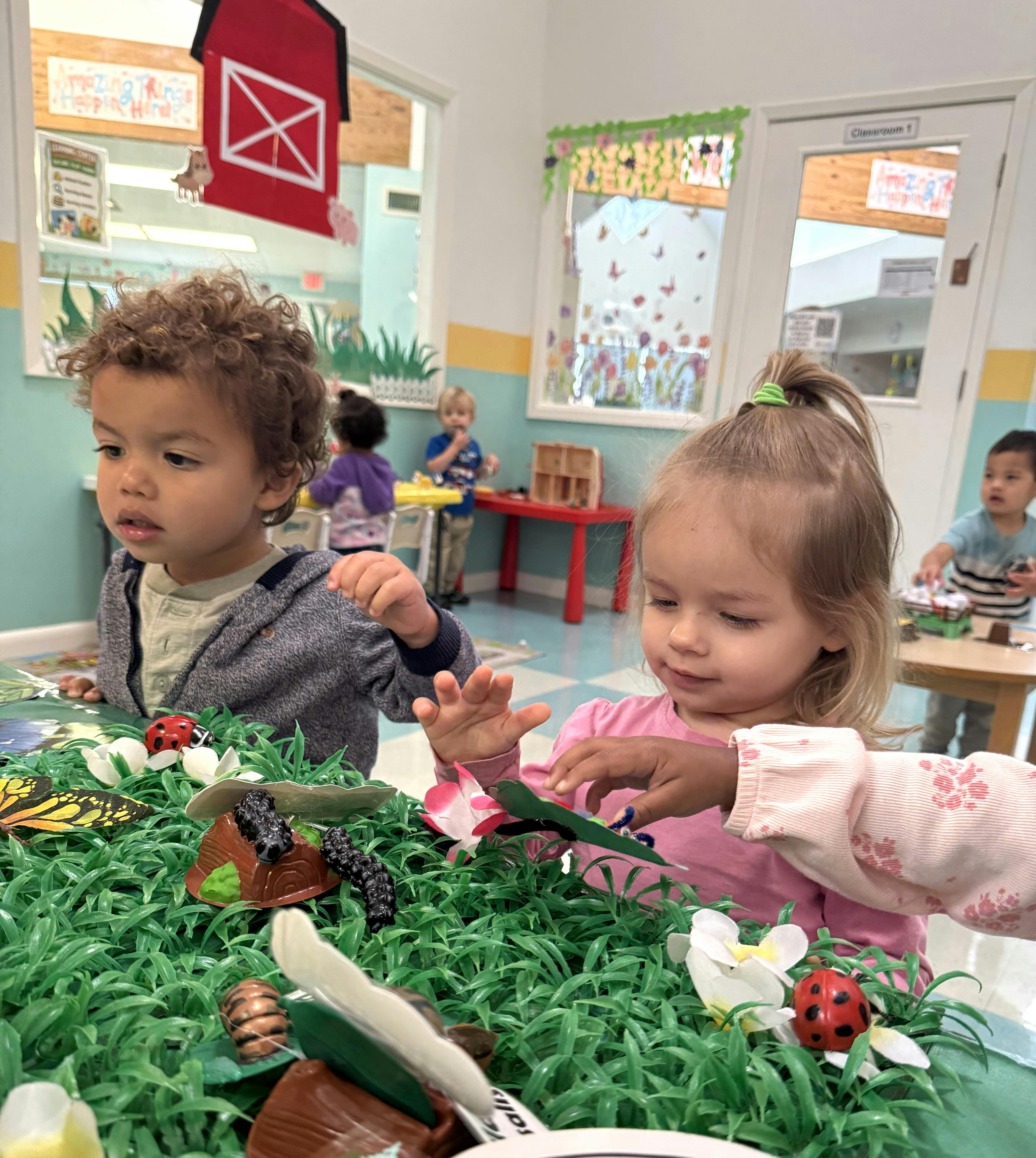 Two children playing with a sensory bin containing artificial grass and insects