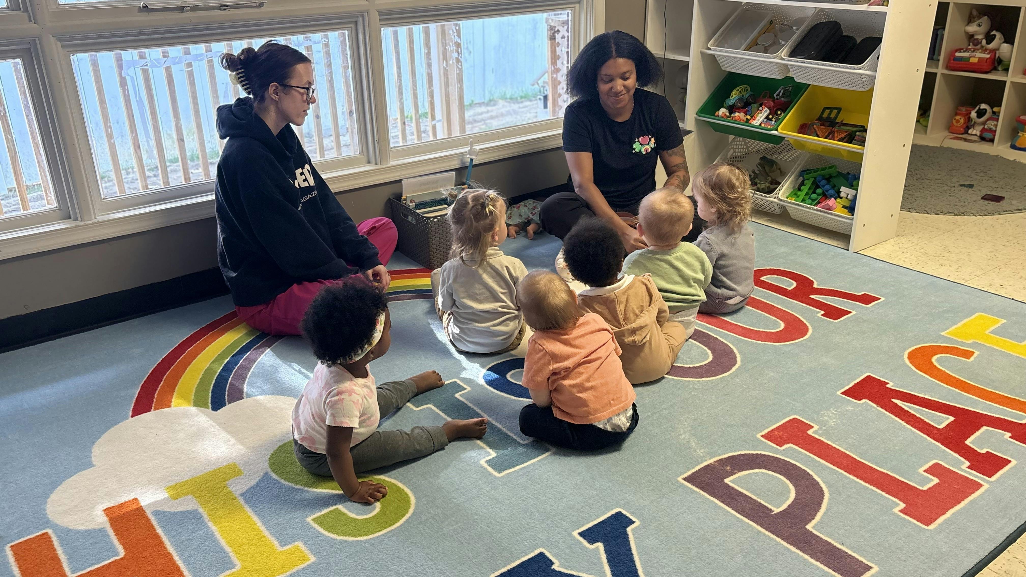 Two people sitting on a rainbow rug with five babies in a classroom