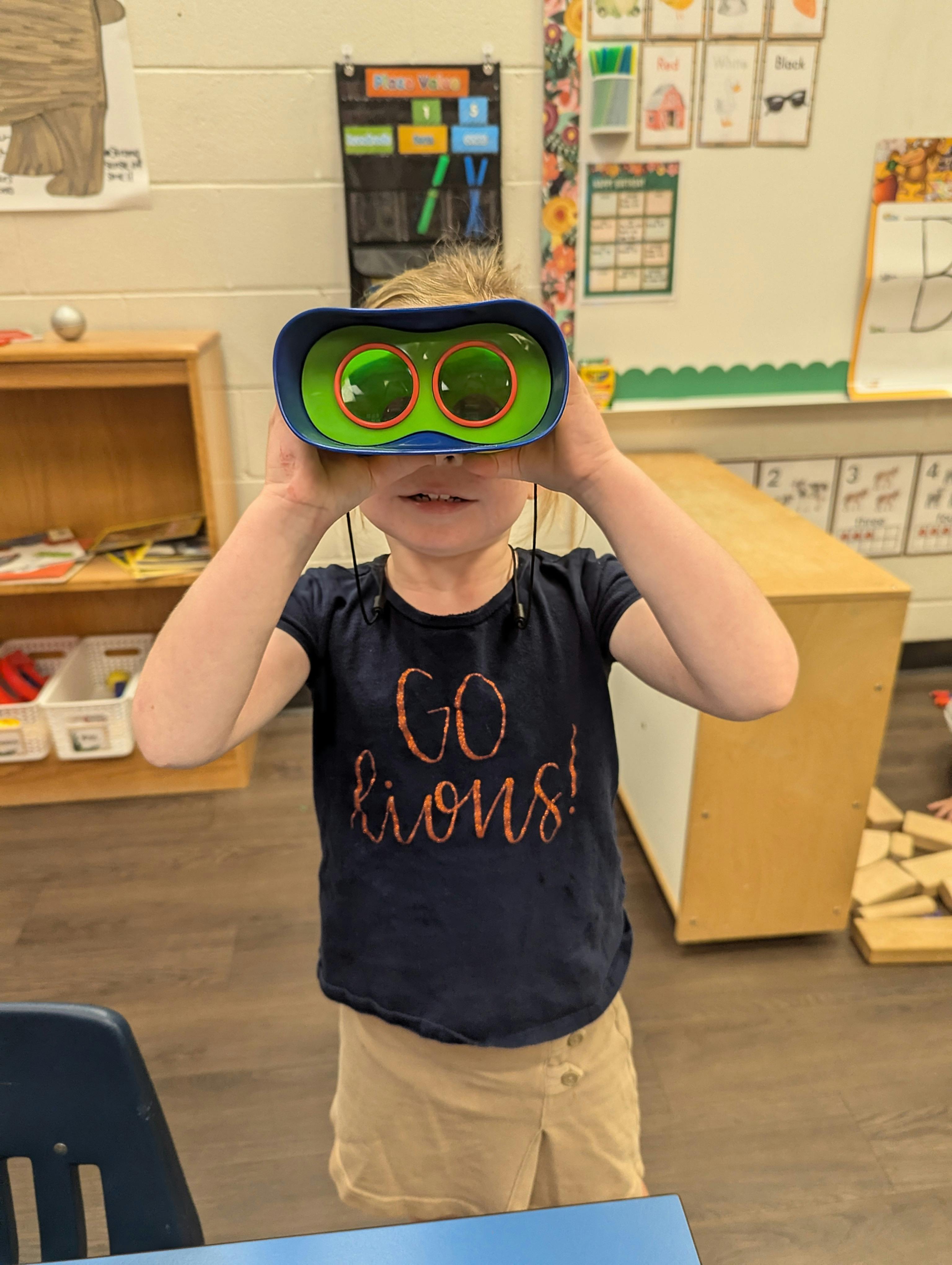 Child holding toy binoculars to their eyes in a classroom