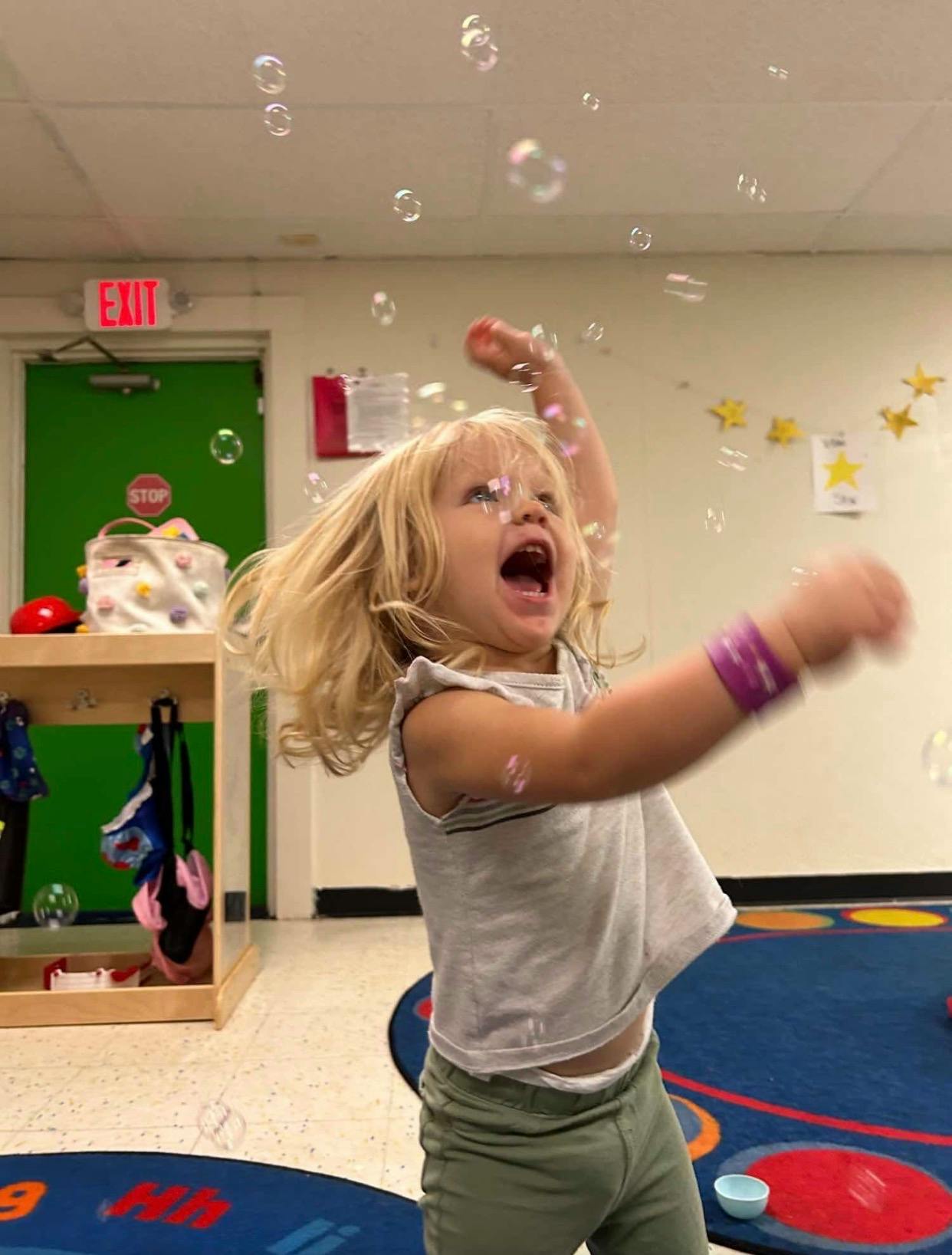 Child with long blonde hair reaching for bubbles in a classroom