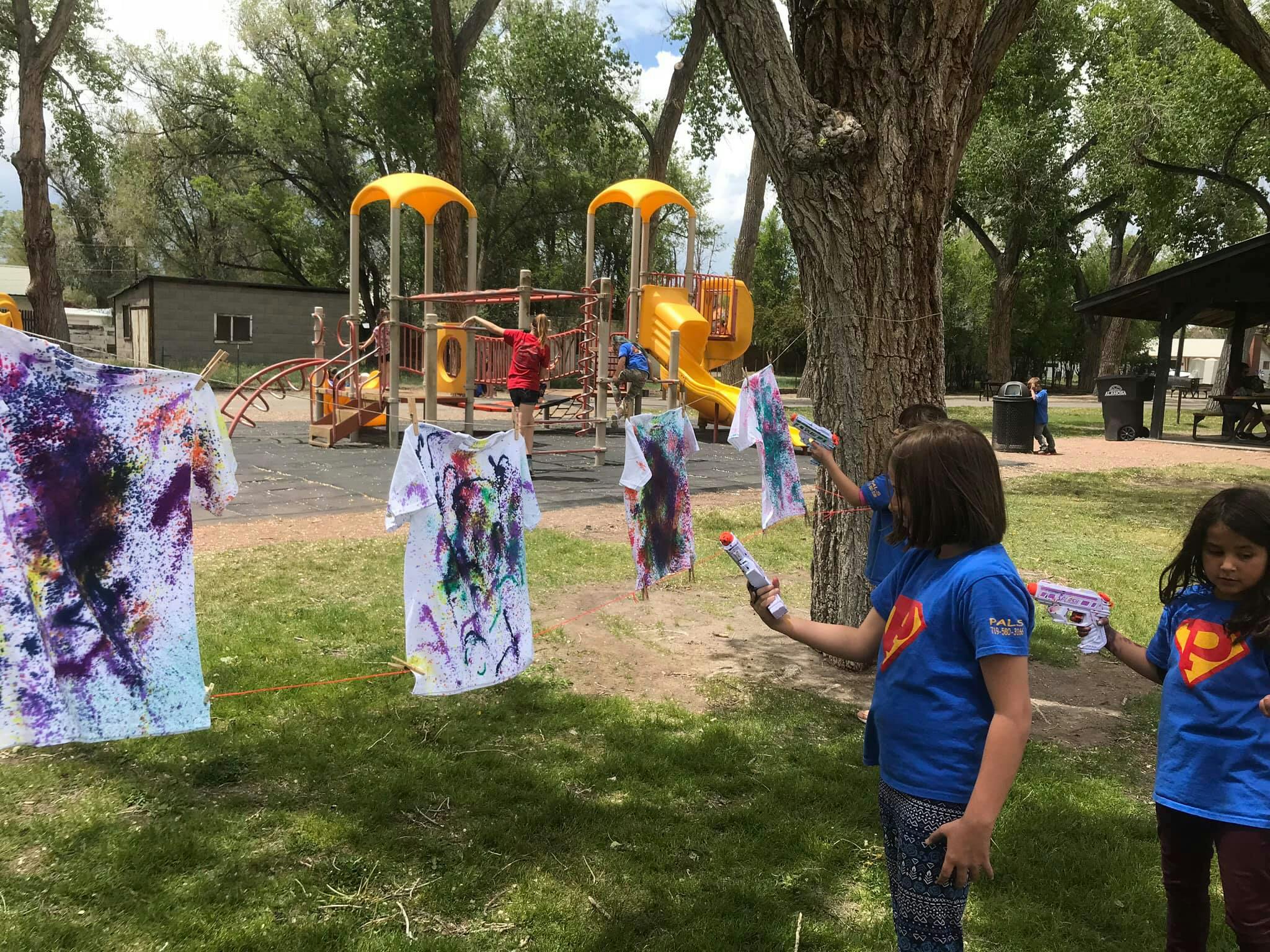 Children painting shirts with squirt guns outdoors near a playground