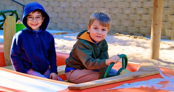 Two children playing in a sandbox, one holding a steering wheel