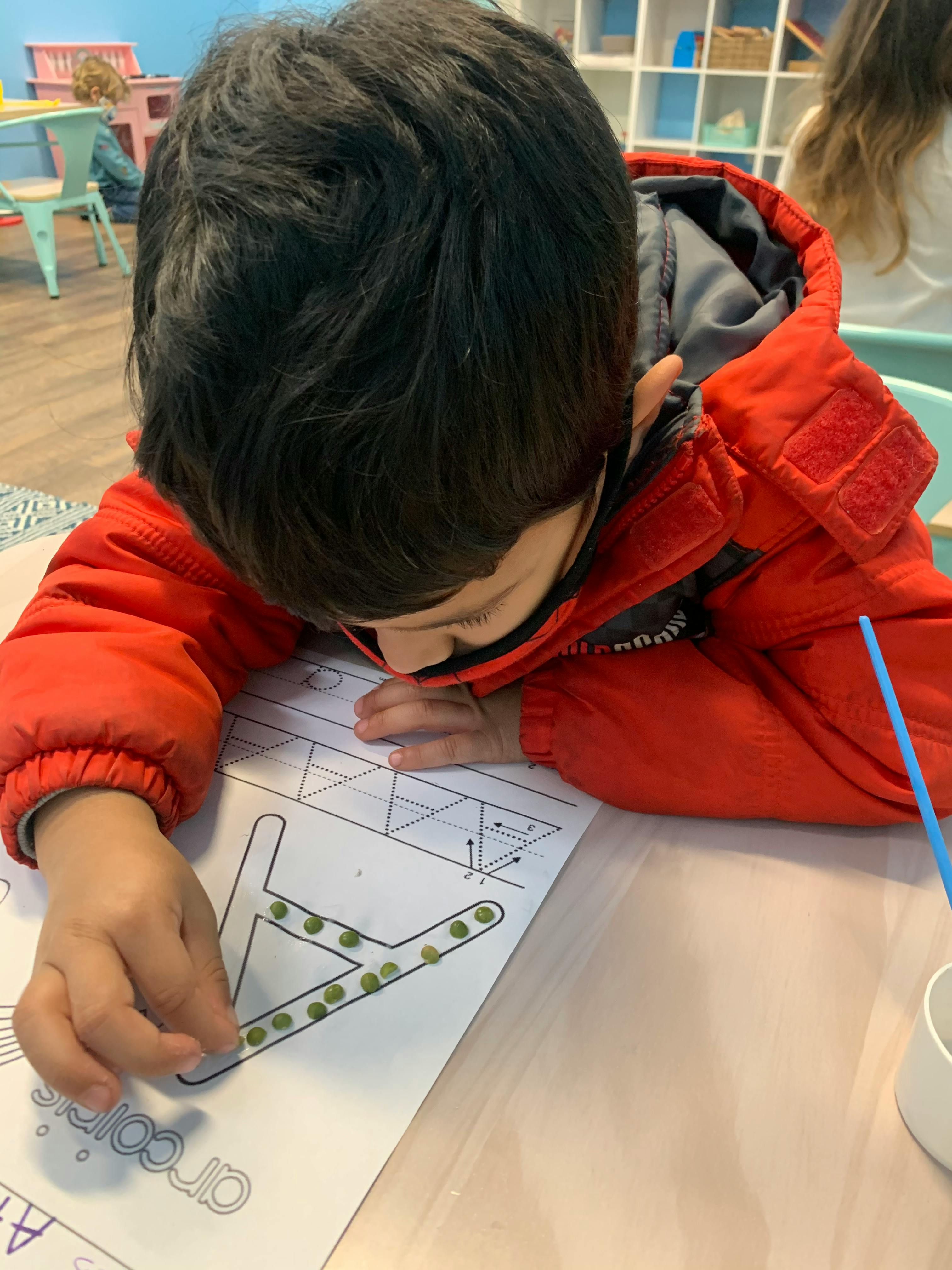 Child in red jacket working on a paper with a letter outline