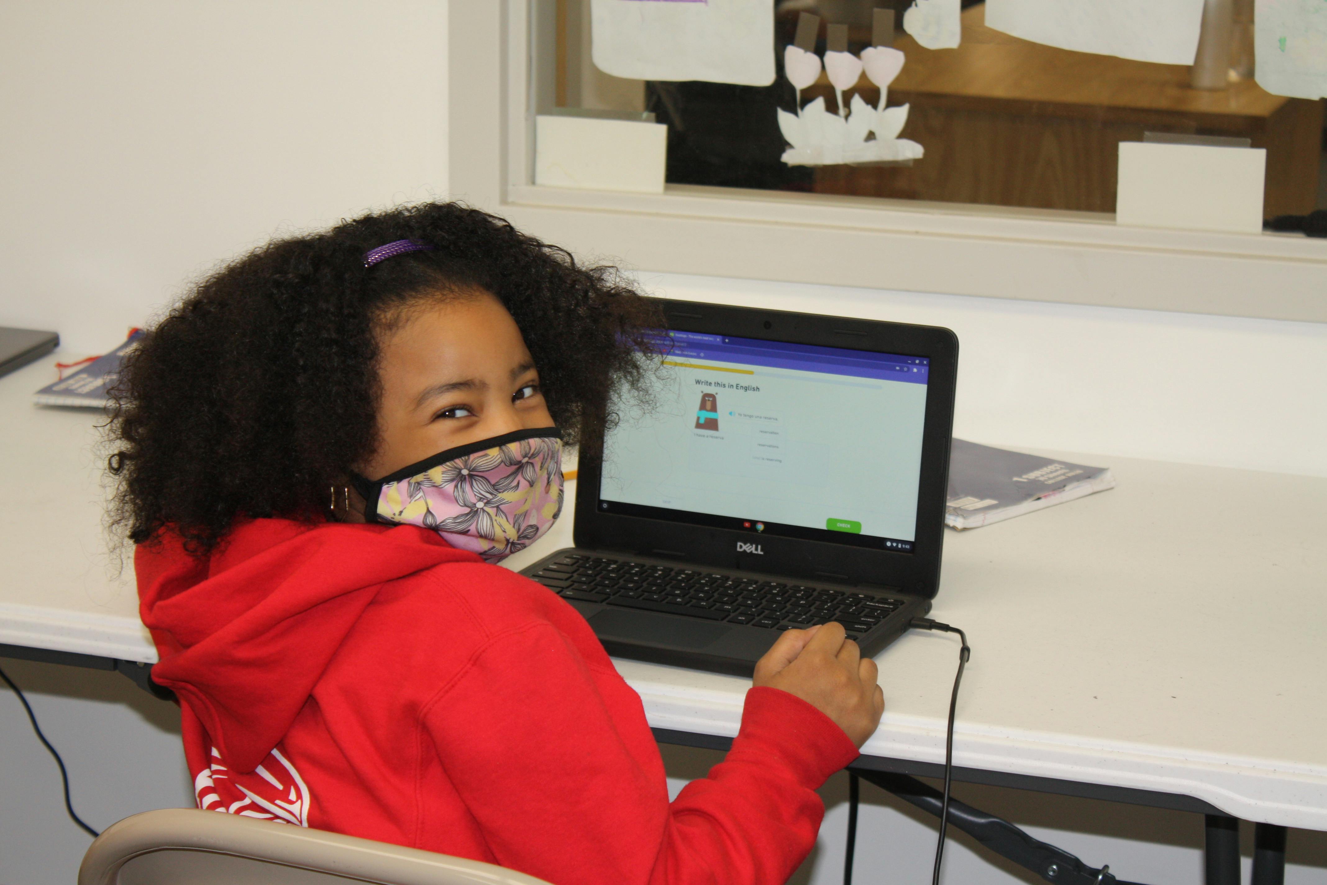 Child in a mask sitting at a desk with a laptop