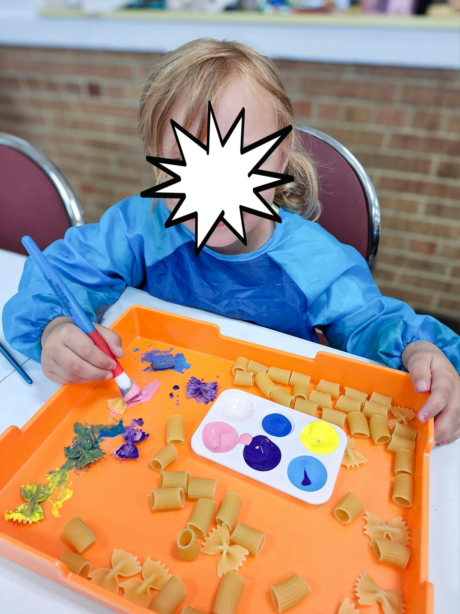Child painting pasta shapes on an orange tray