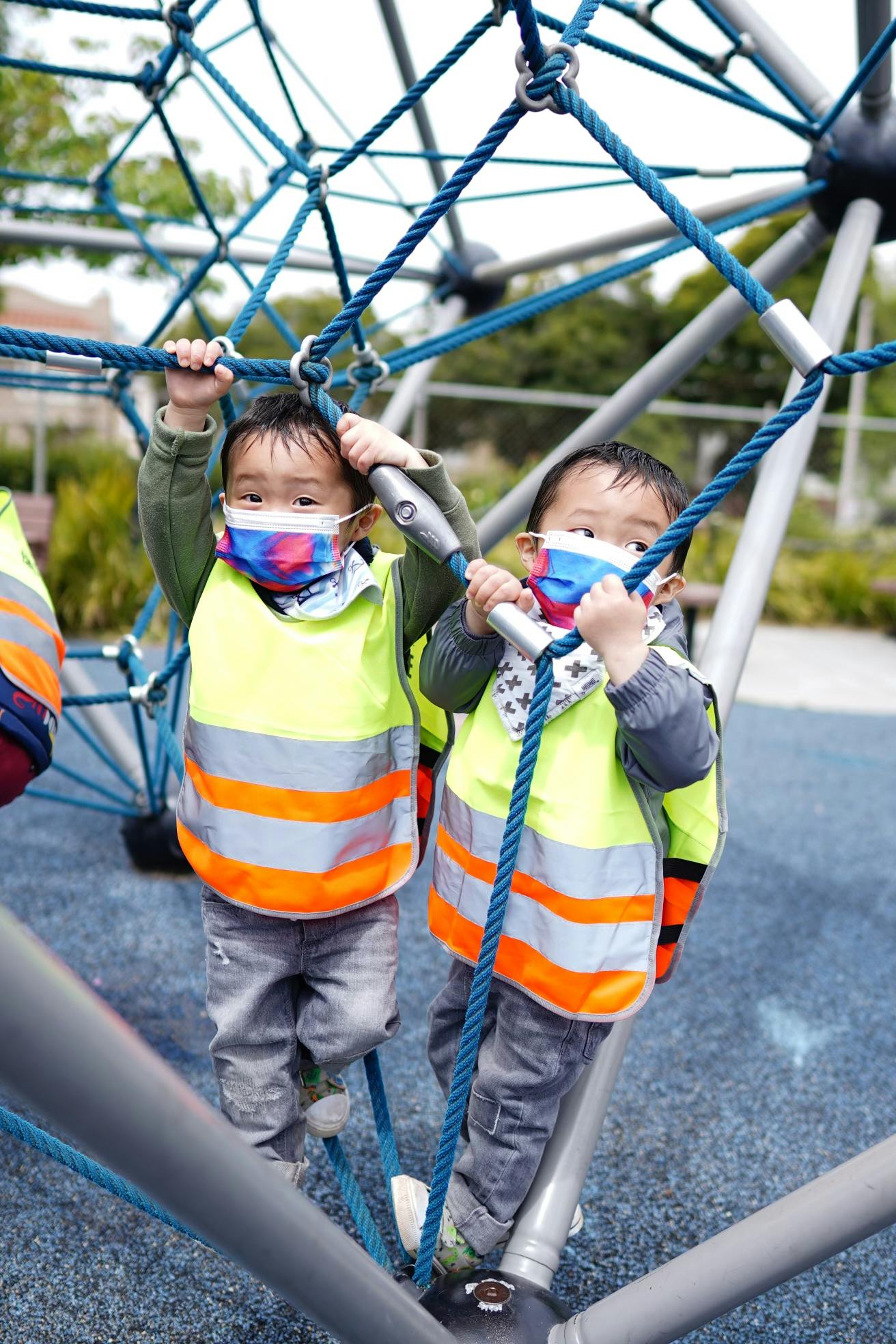 Two children in masks and safety vests climbing on a rope structure