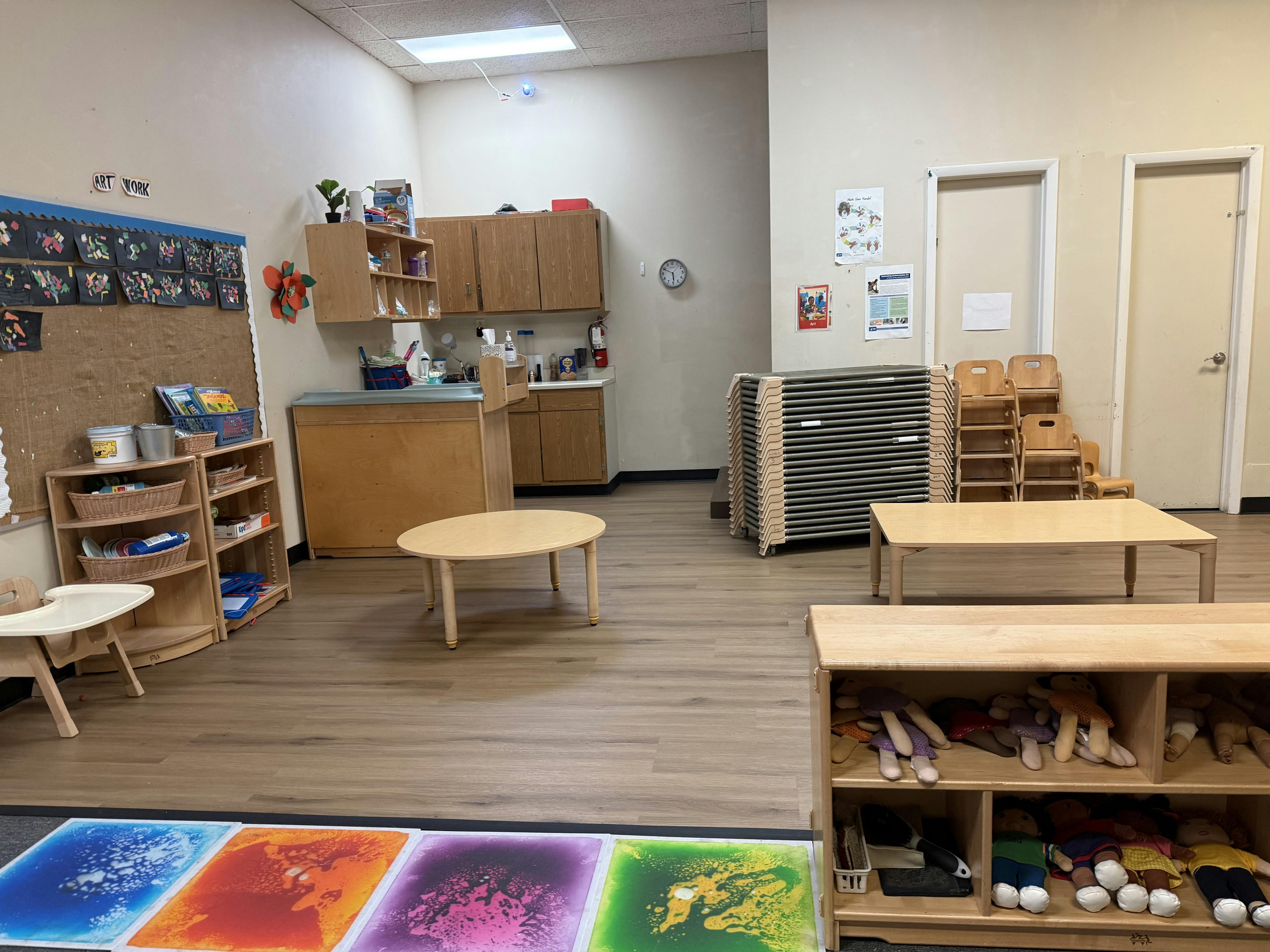 Empty classroom with small tables and chairs, shelves, and a kitchen area