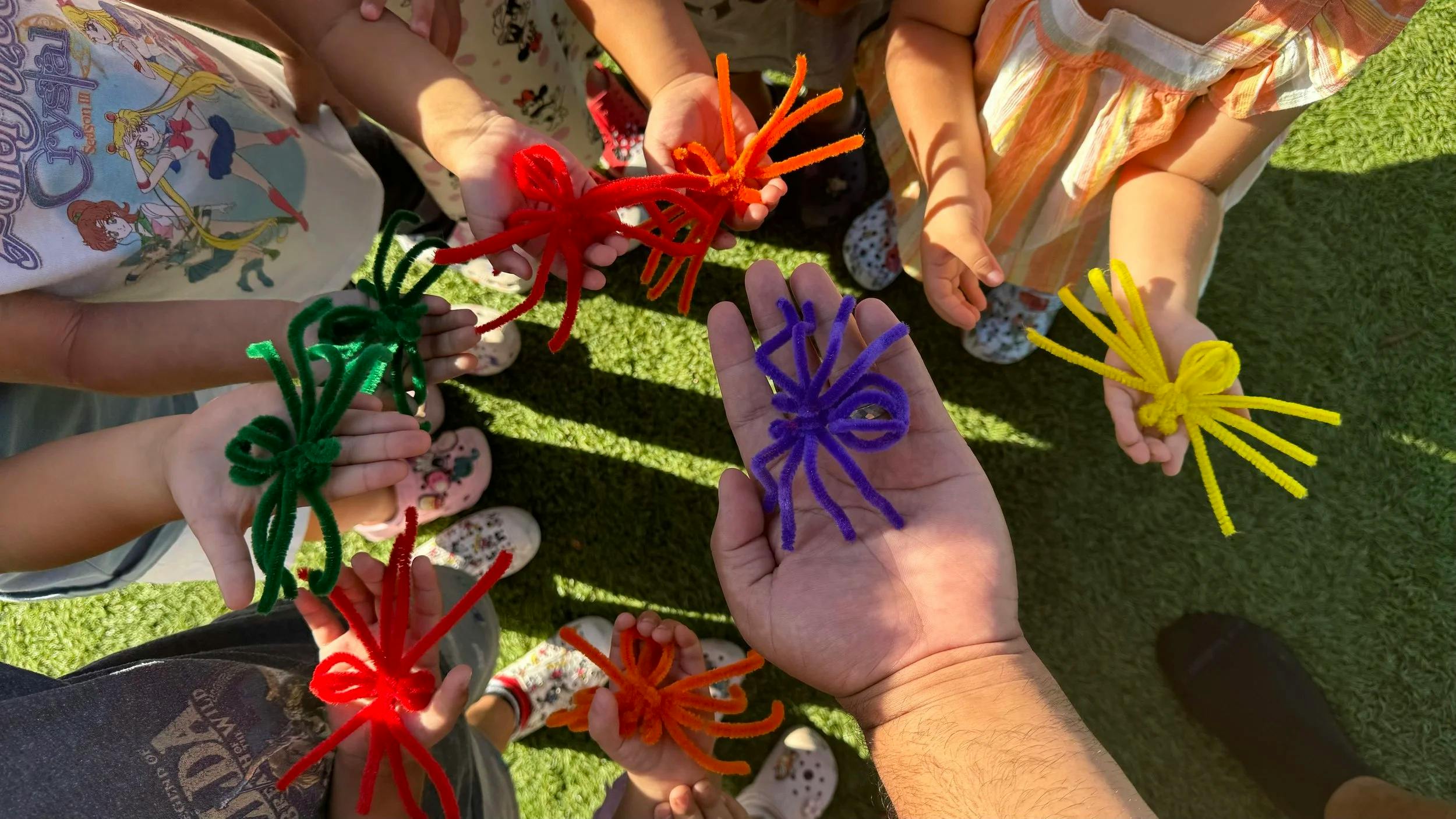 Multiple hands holding pipe cleaner spiders in various colors