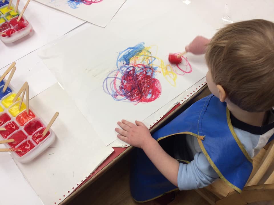 Child in a blue smock painting with red, blue, and yellow paint