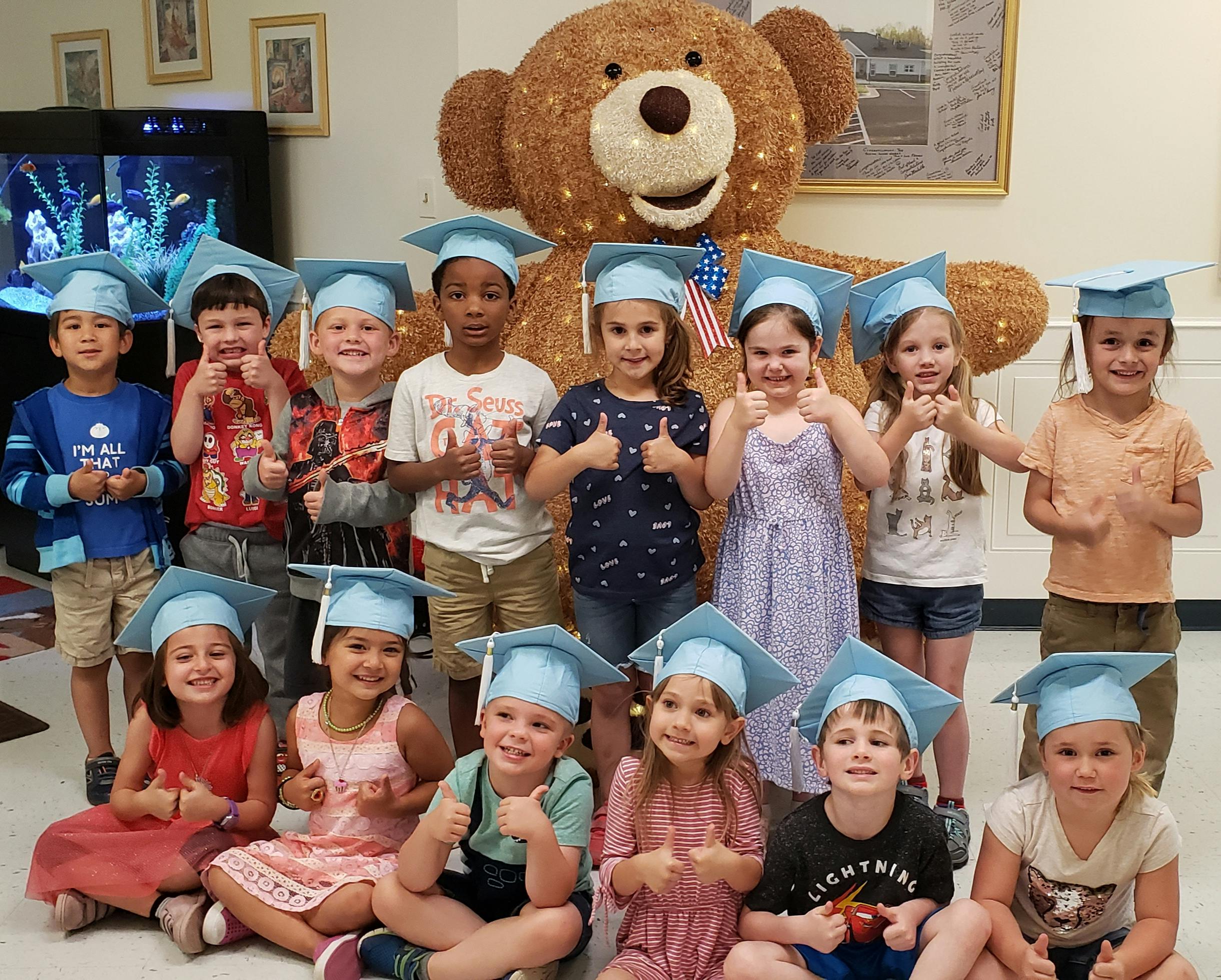 Group of children wearing graduation caps, some giving thumbs up, with a large teddy bear