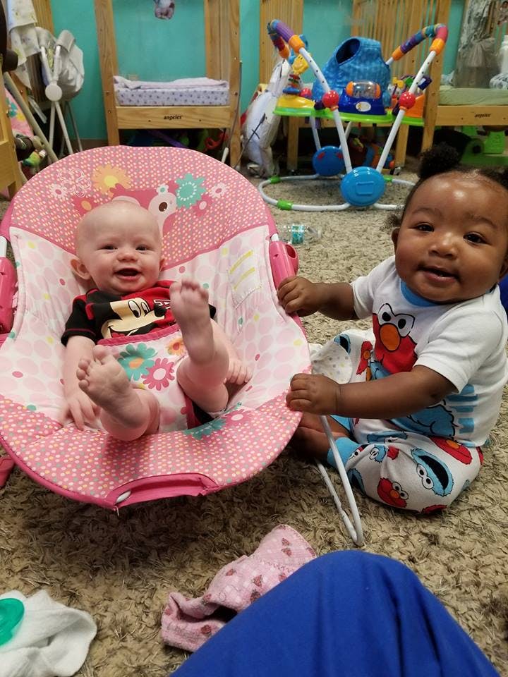 Two babies playing on the floor, one in a pink bouncer