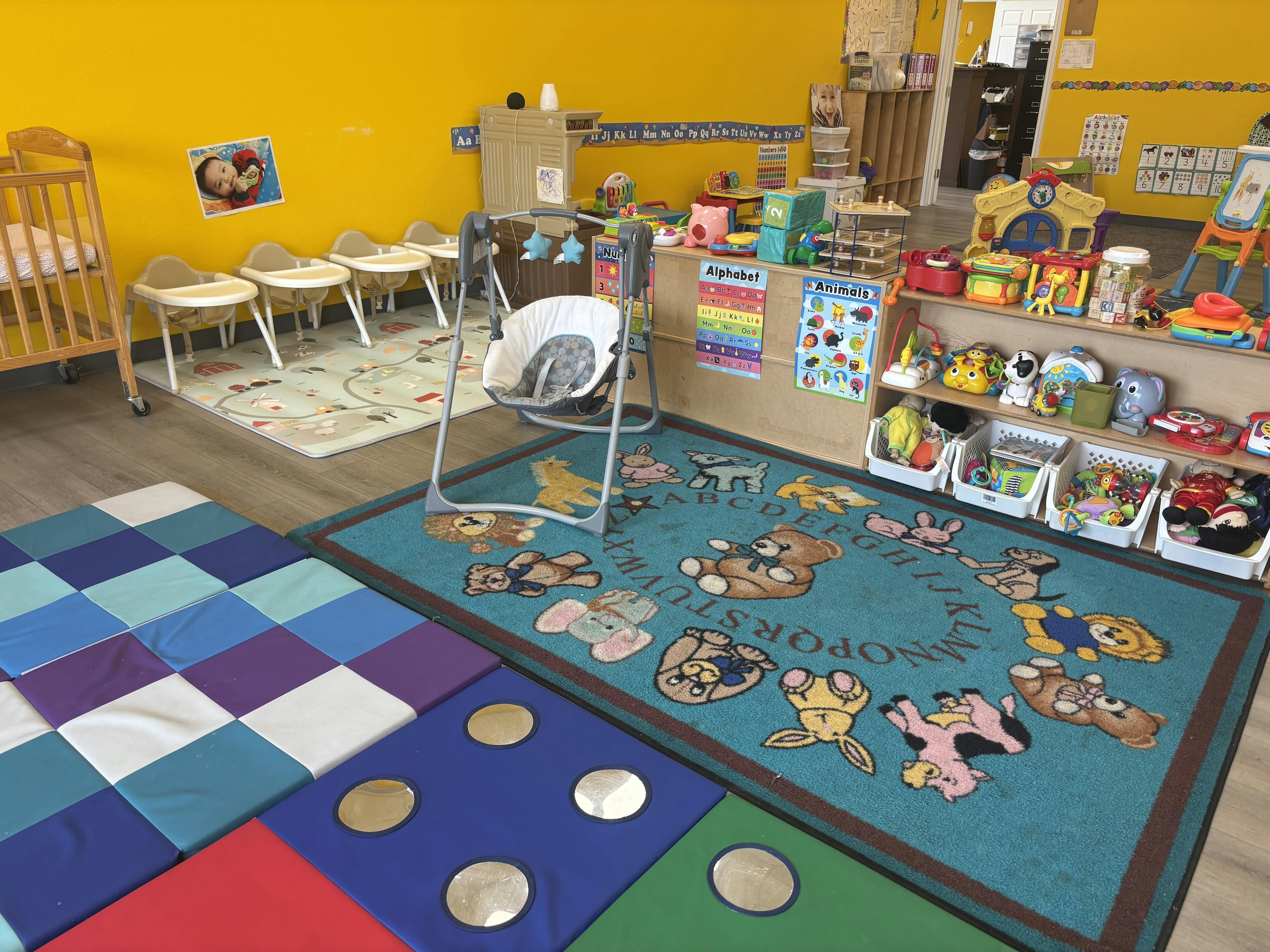 Brightly colored daycare room with play rugs, infant seats, and various toys