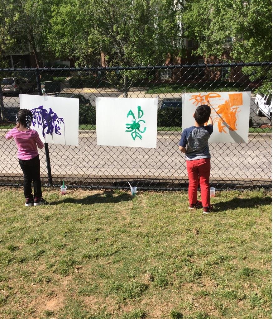 Two children painting on paper attached to a fence outdoors