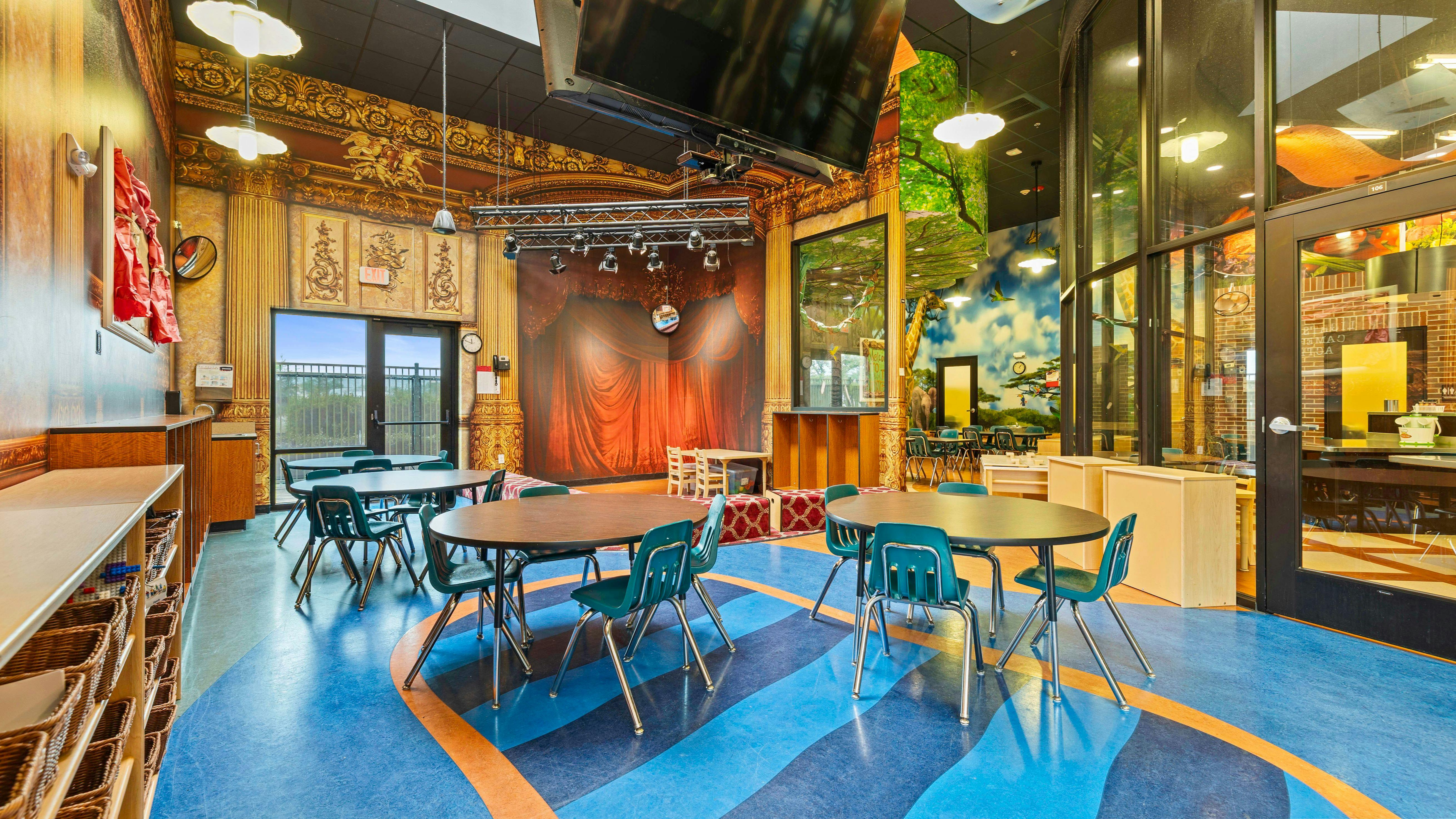 Classroom with tables and chairs, a stage, and murals of nature scenes