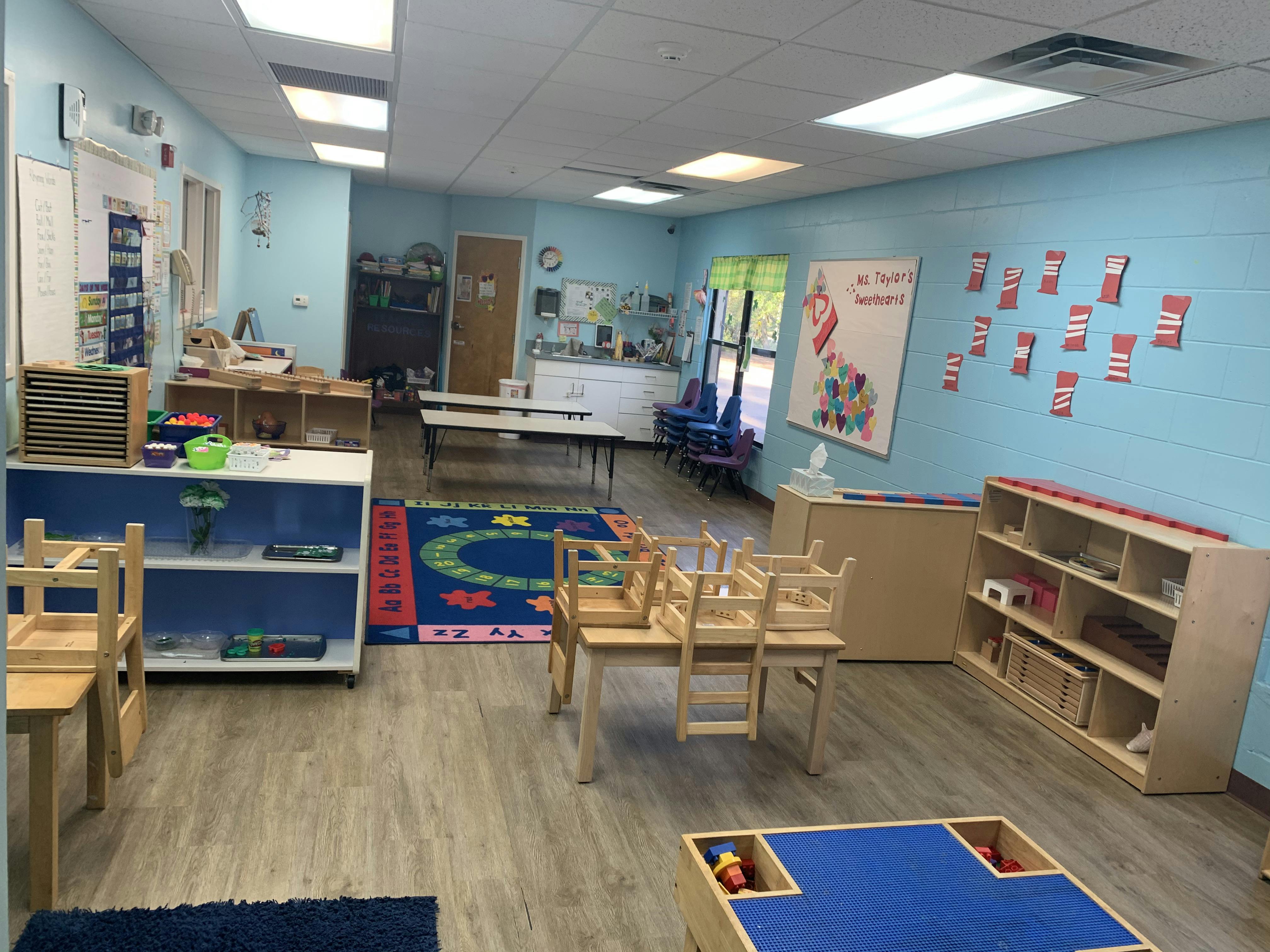 Classroom with tables, chairs, shelving units with materials, and a colorful rug