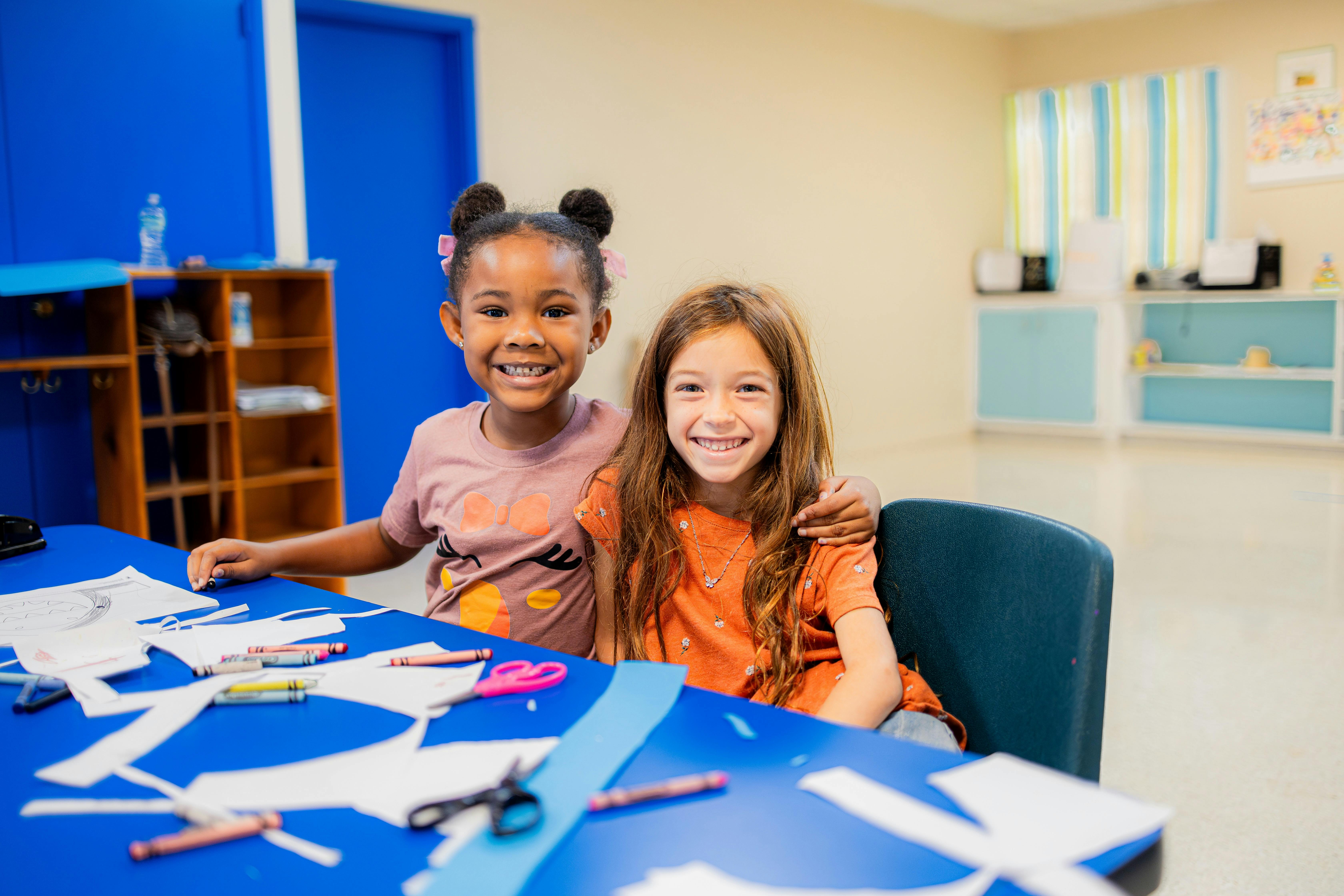 Two children sitting at a table with paper and art supplies in a classroom