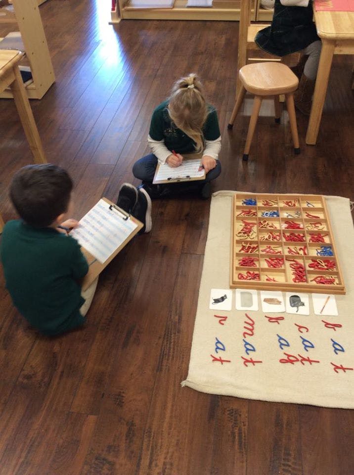 Two children writing on clipboards on a wooden floor next to a learning mat