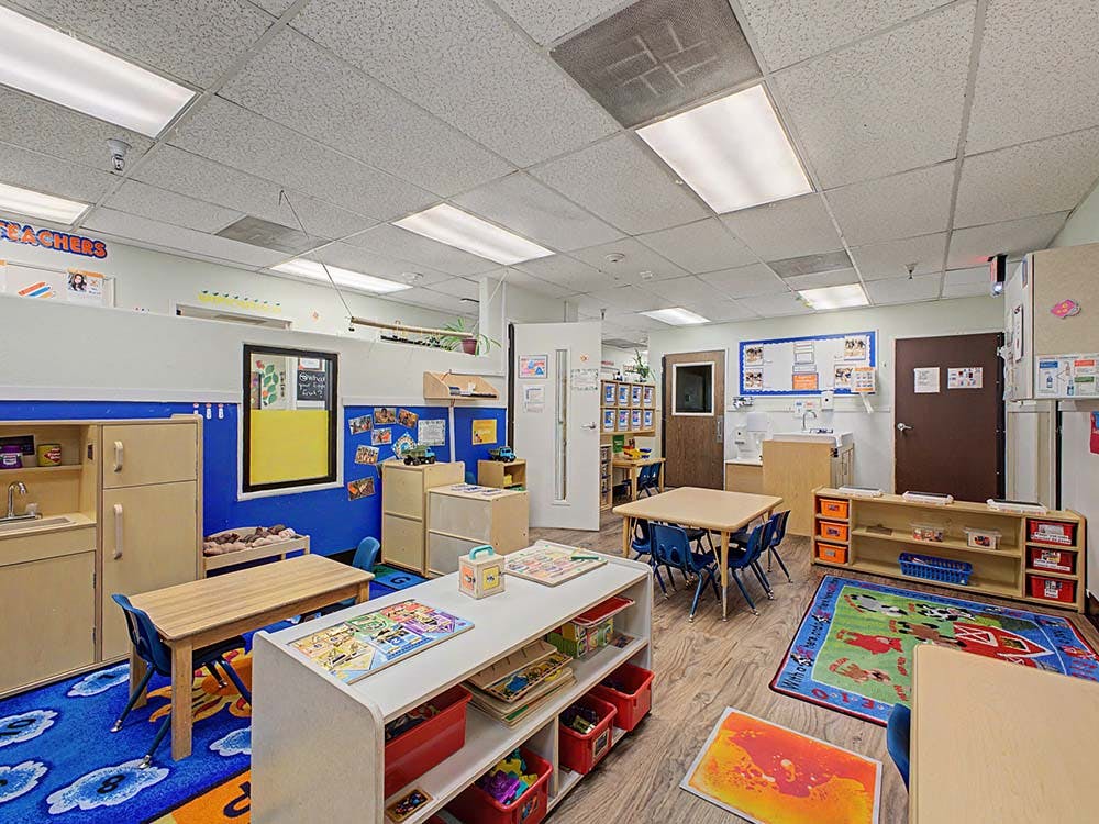 An empty classroom with tables, chairs, shelving units, and a small play kitchen