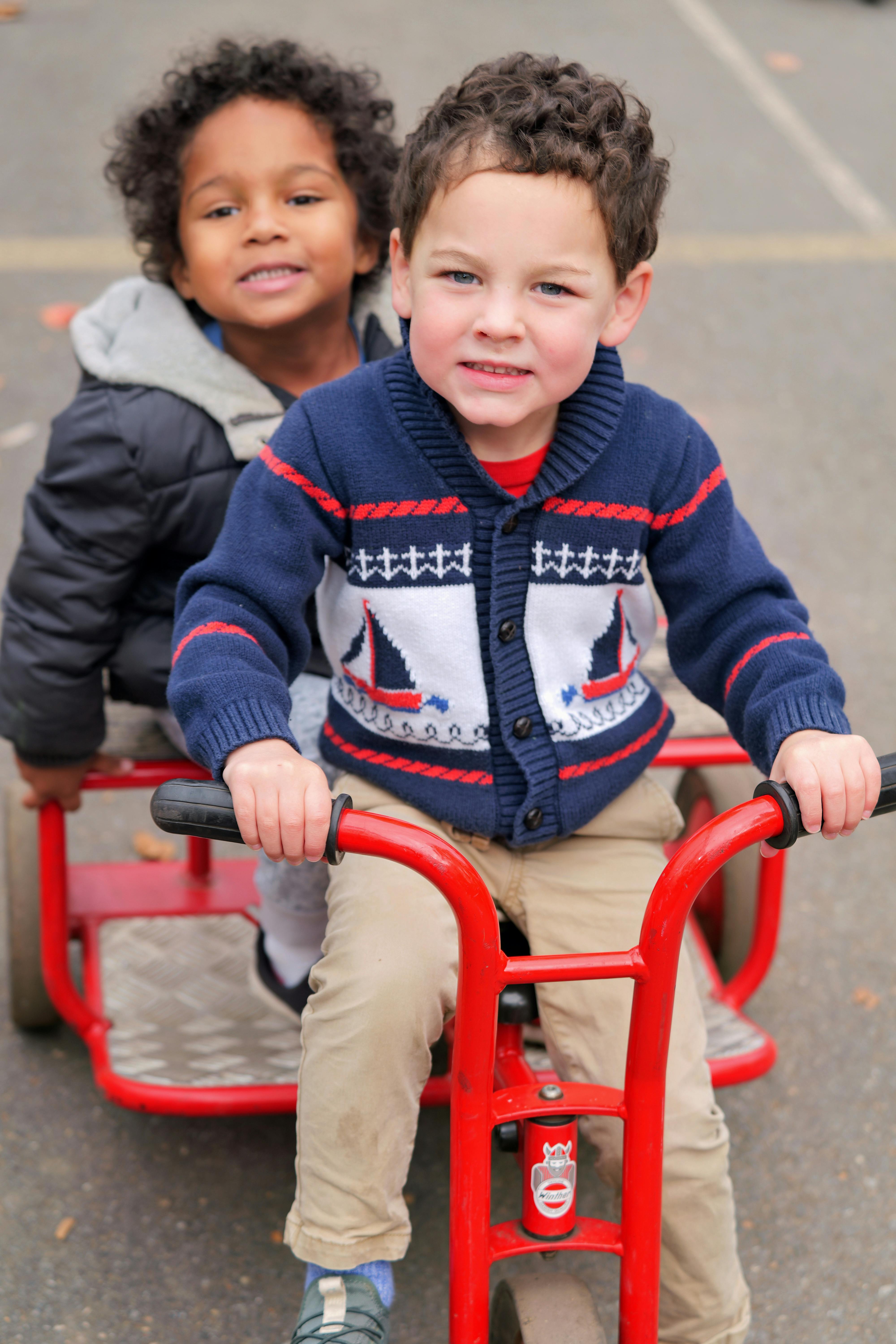 Two children riding a red tricycle together on a paved playground