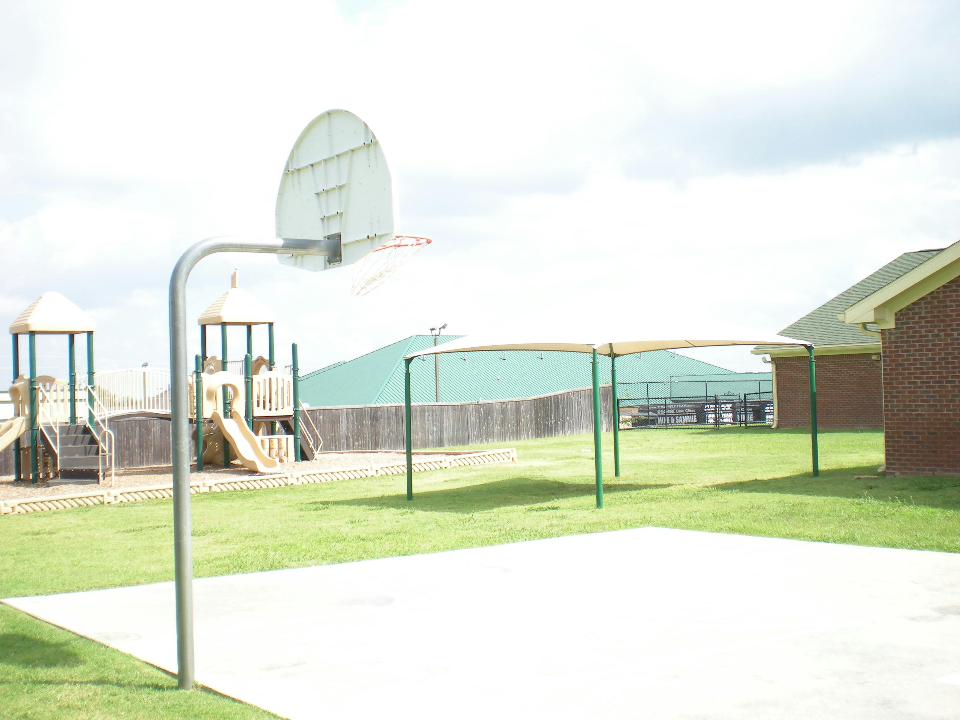 Basketball hoop on a court with a playground and shade structure in the background