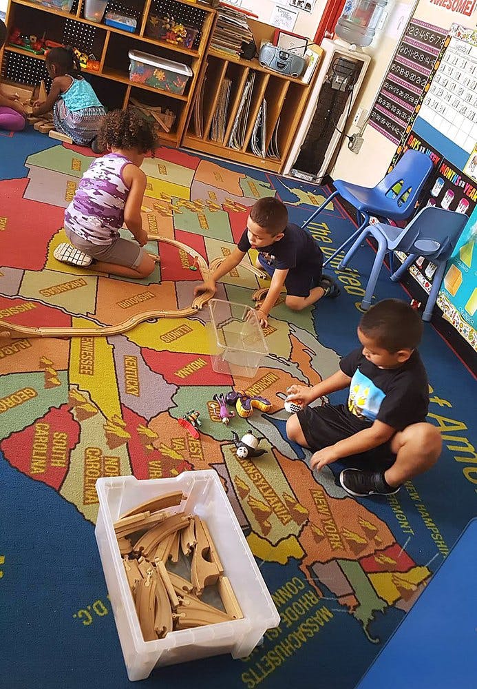 Three children playing with toy trains on a map rug in a classroom