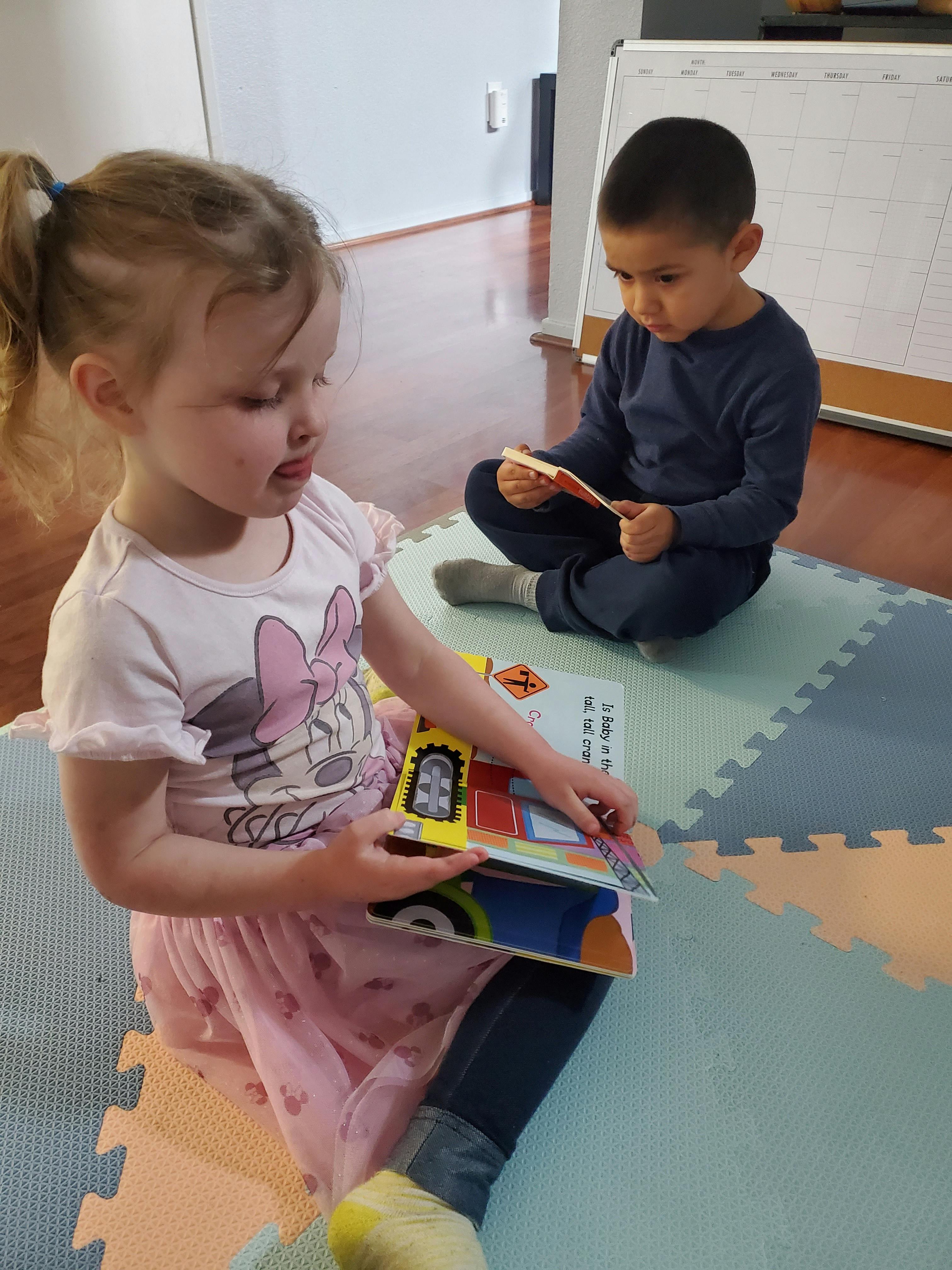 Two children sitting on a mat, one reading a book and the other looking at a book