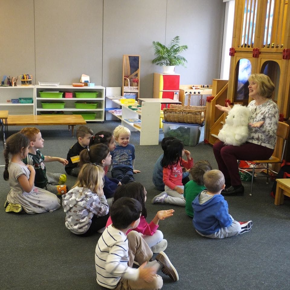 Adult holding a white stuffed animal, speaking to a group of children