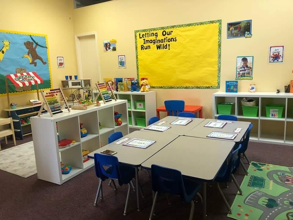 Classroom with tables and chairs, shelves with toys, and a yellow bulletin board