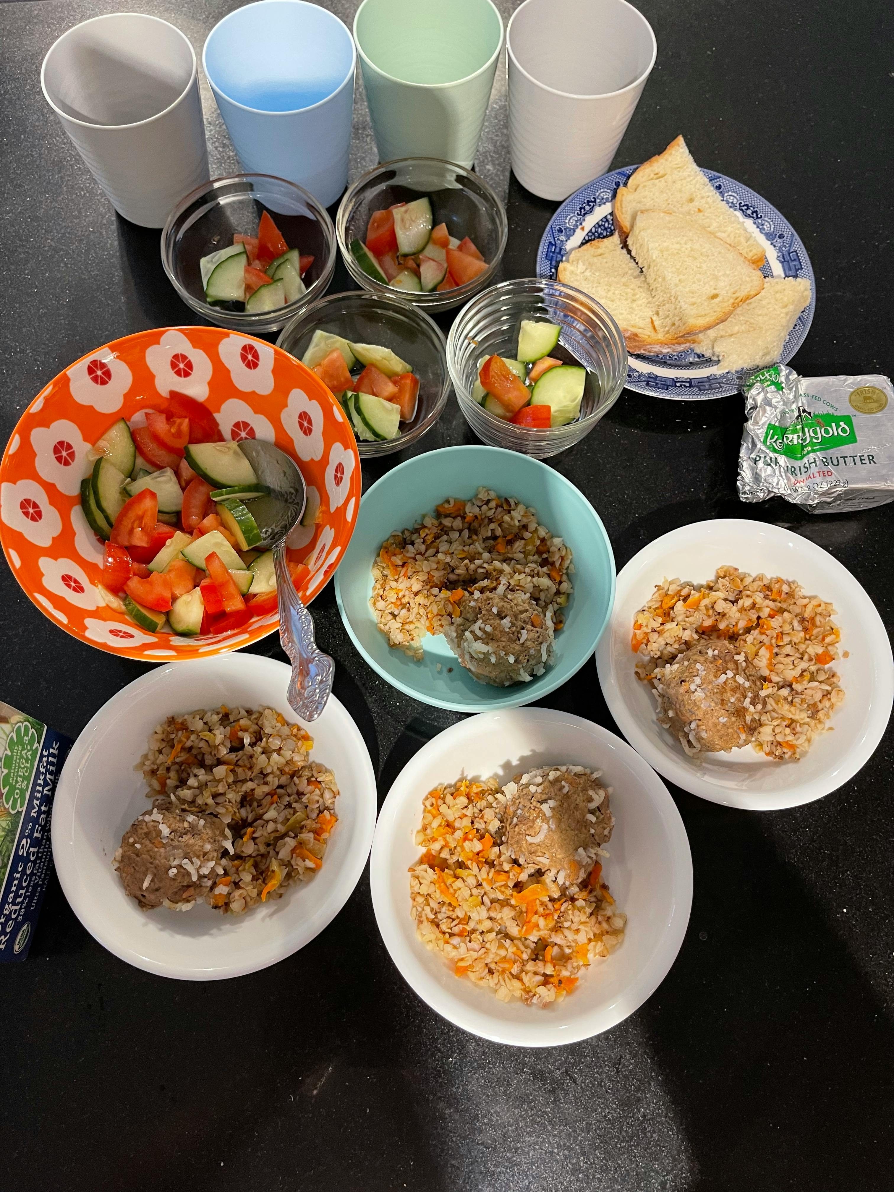 Multiple bowls of food including rice, meat, and salad, with cups and bread