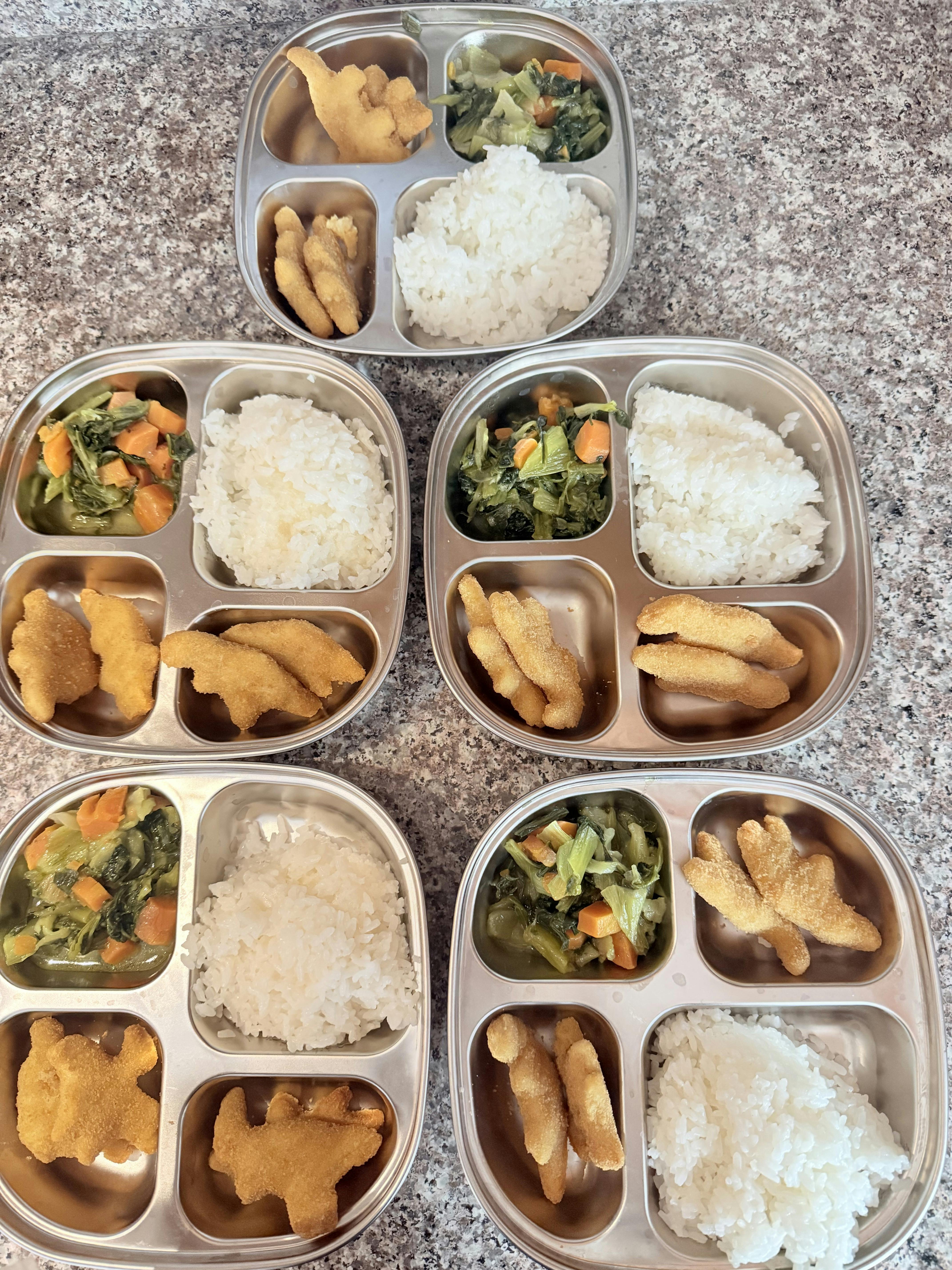 Five stainless steel divided trays containing rice, cooked vegetables, and nuggets