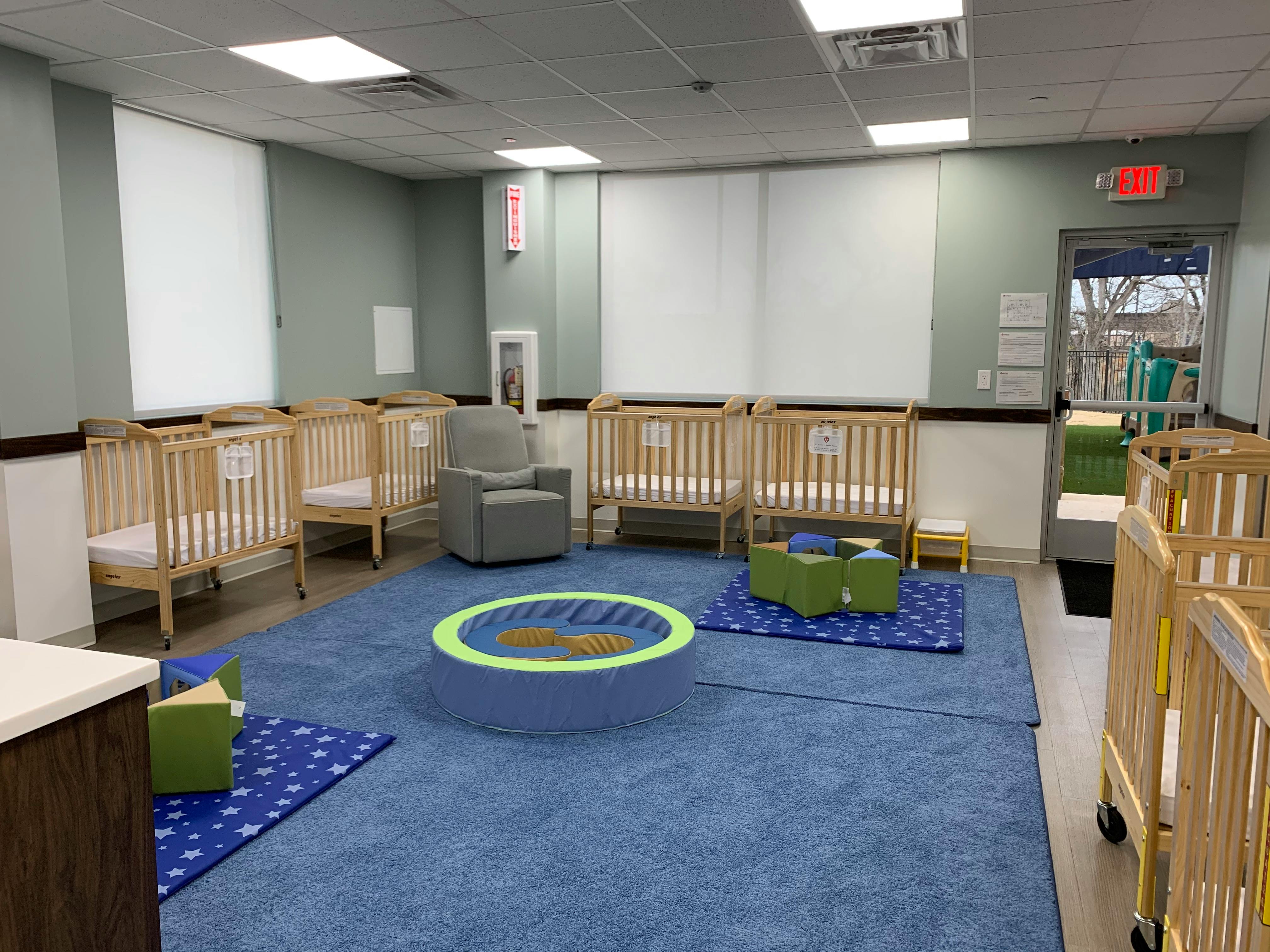 Room with multiple cribs, a chair, and play mats on a blue carpet