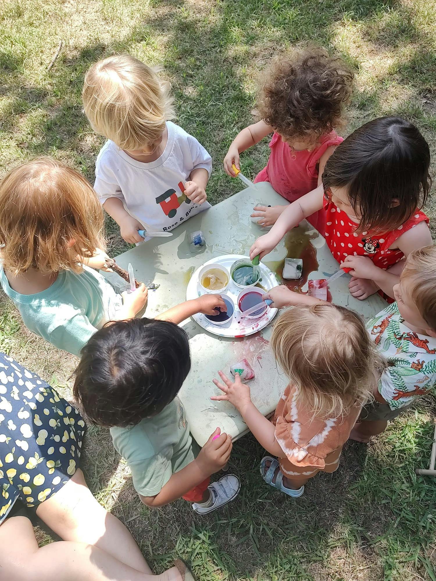 Children gathered around a table painting outdoors, painting with various colors