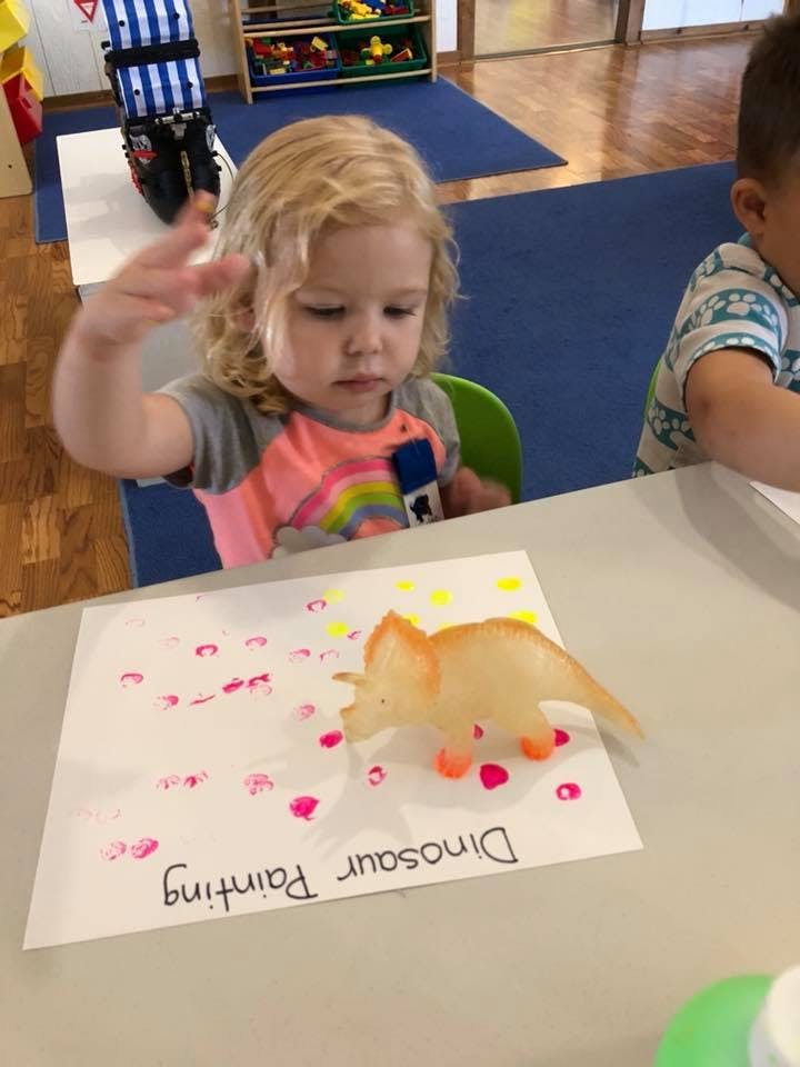 Child at a table with a dinosaur toy and a paper with "Dinosaur Painting"
