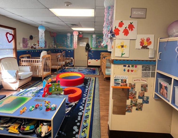 A classroom with cribs, play mats, and child artwork on the wall